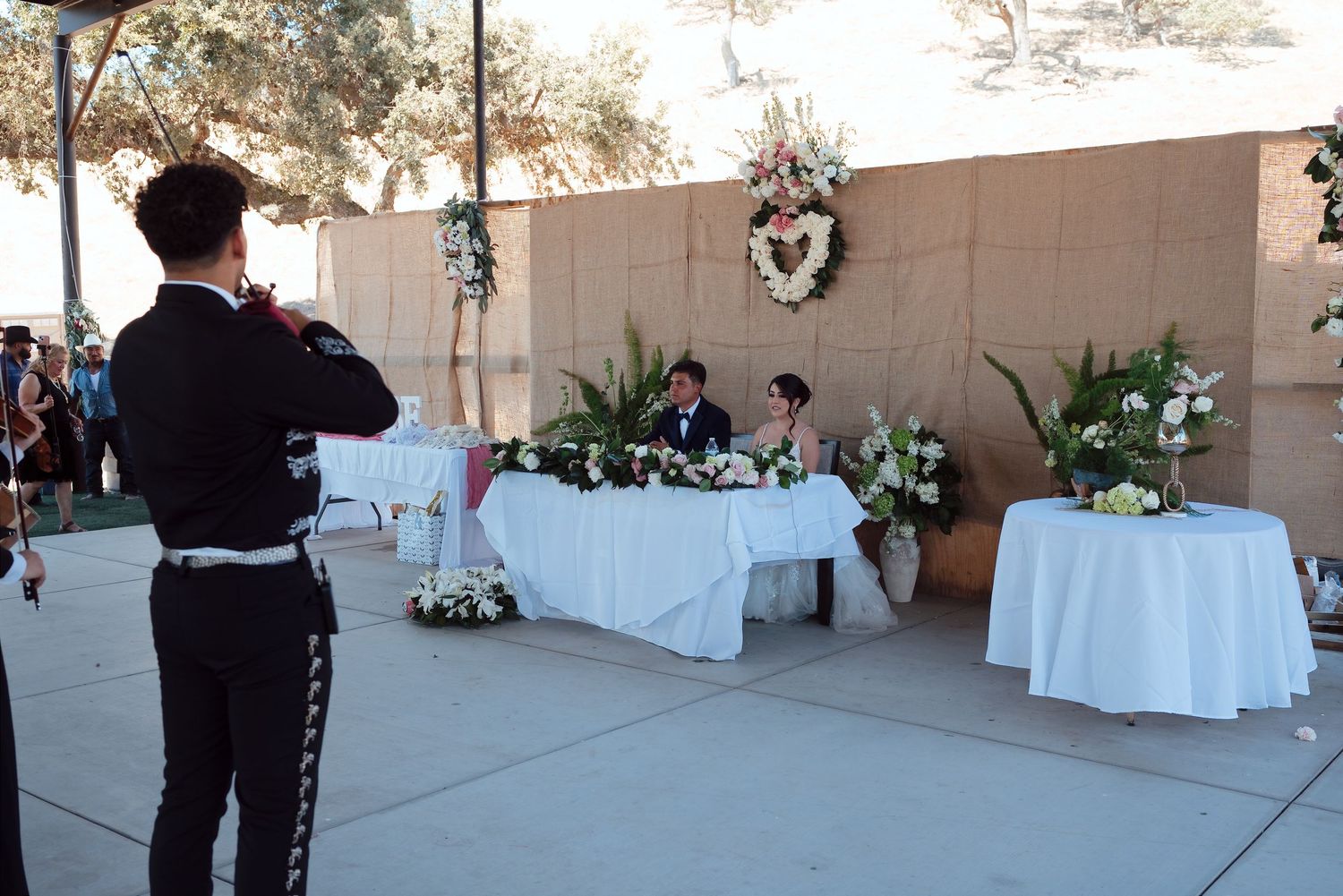 Mariachi performer serenades couple at outdoor wedding ceremony table decorated with white flowers.