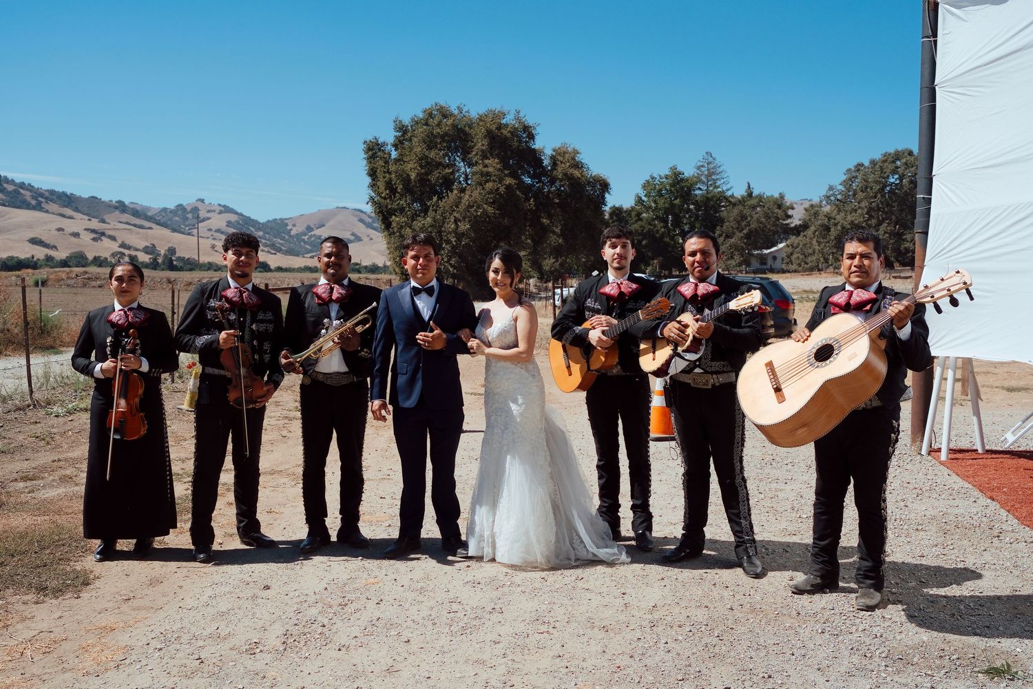 A bride and groom pose with a mariachi band outdoors in a scenic California valley setting.