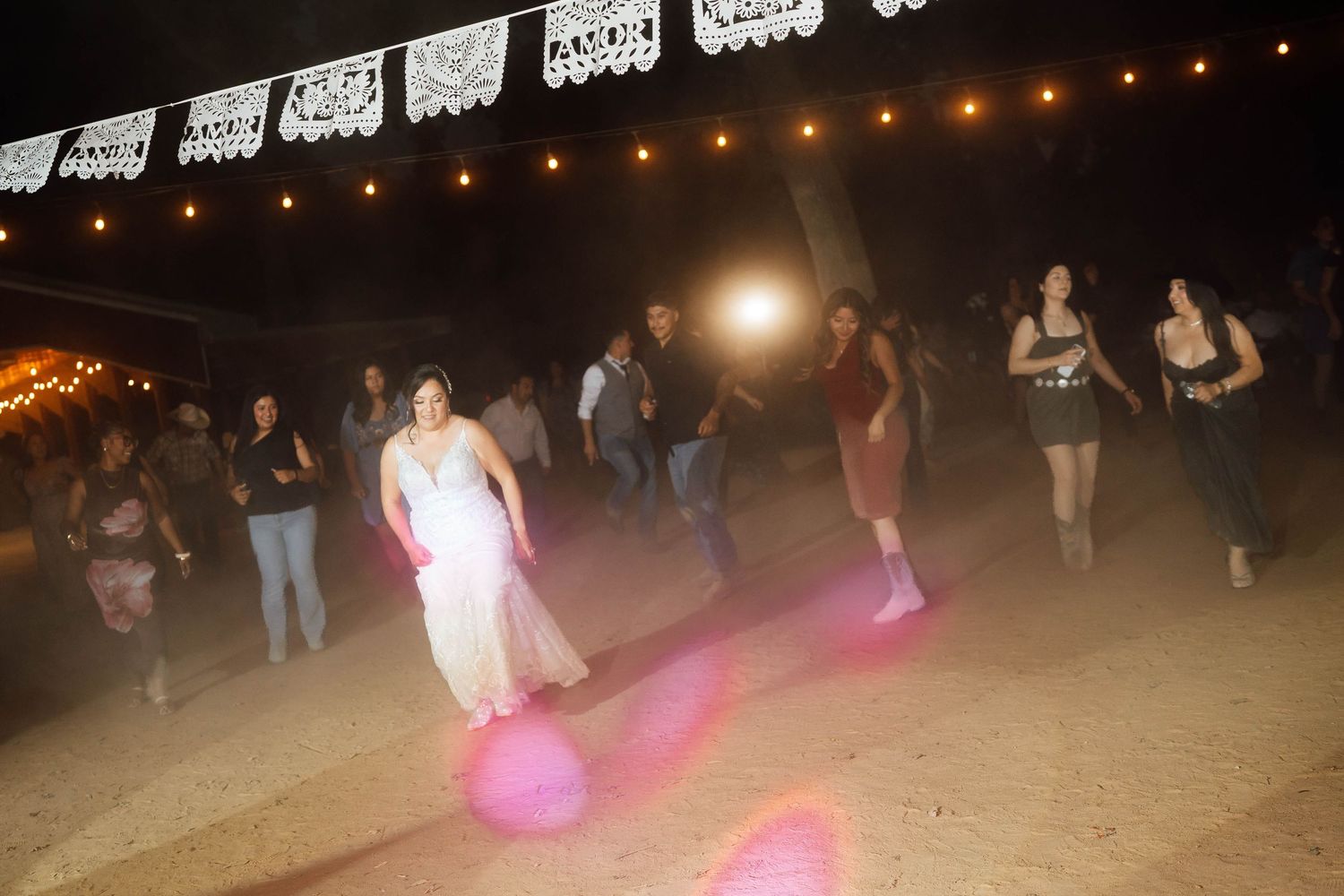 Bride in white gown dancing with guests on outdoor dirt dance floor at night with string lights.