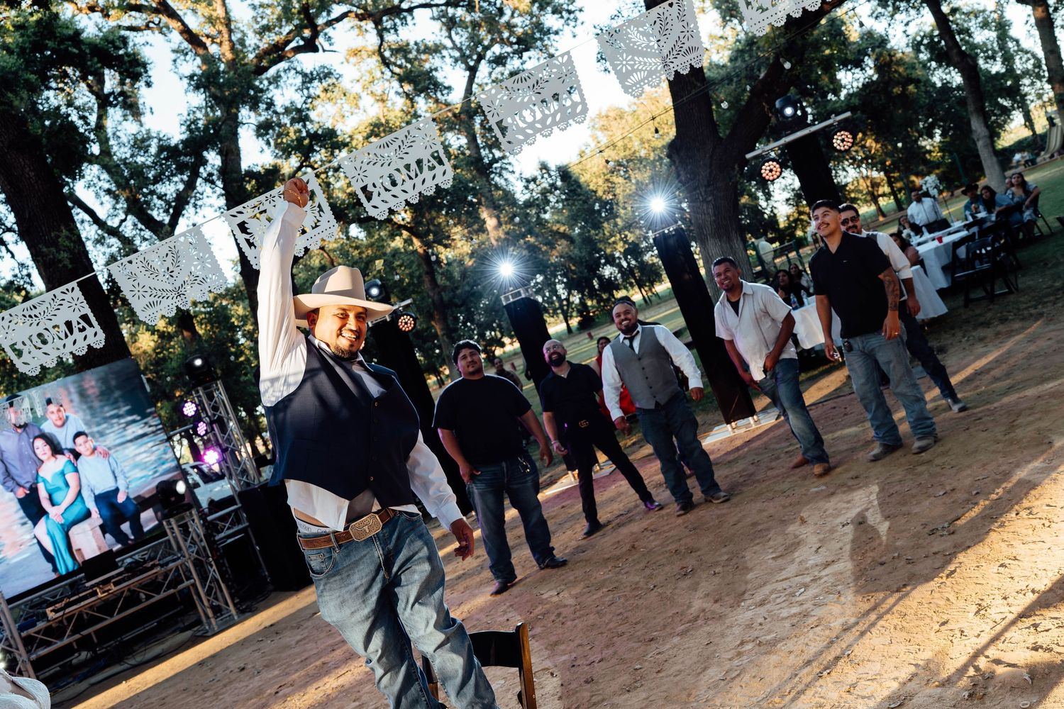 Group of men in cowboy hats dancing at an outdoor evening wedding reception with stage lights.