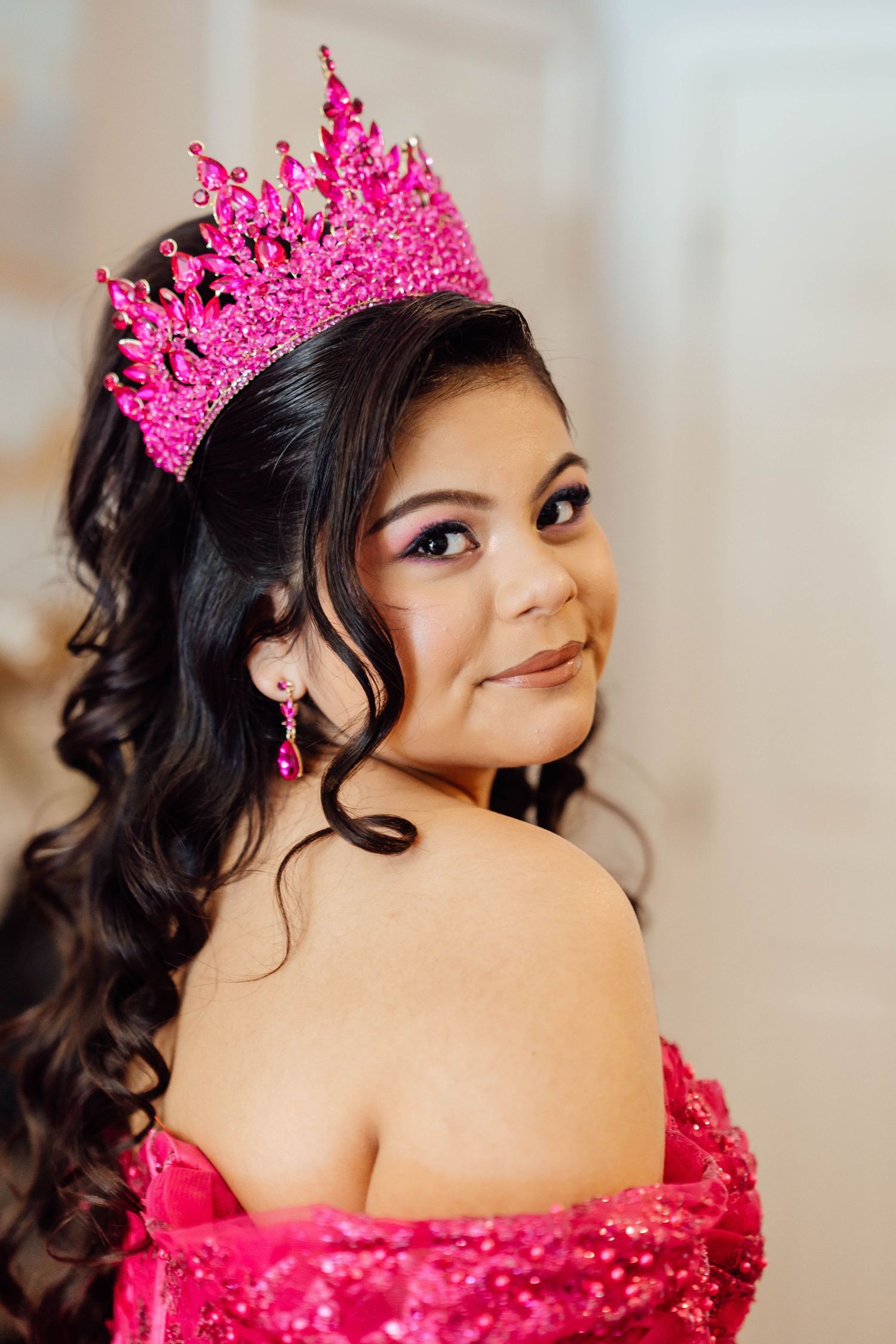 Young Latina girl wearing a pink rhinestone crown and hot pink quinceañera gown smiling over shoulder.