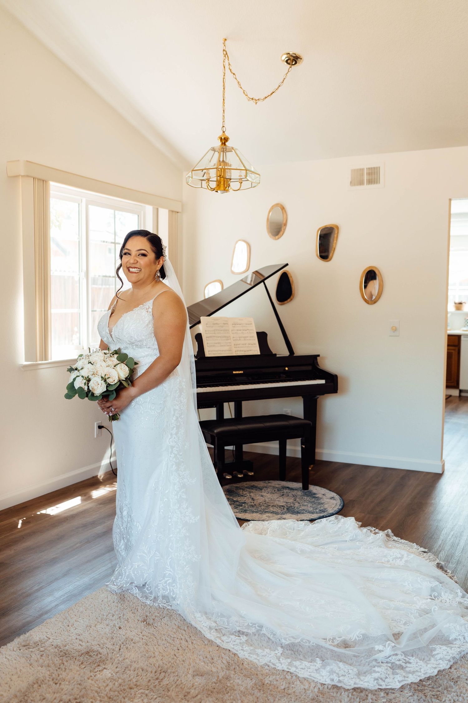 Bride in lace wedding dress with long train posing with white bouquet near black grand piano indoors.