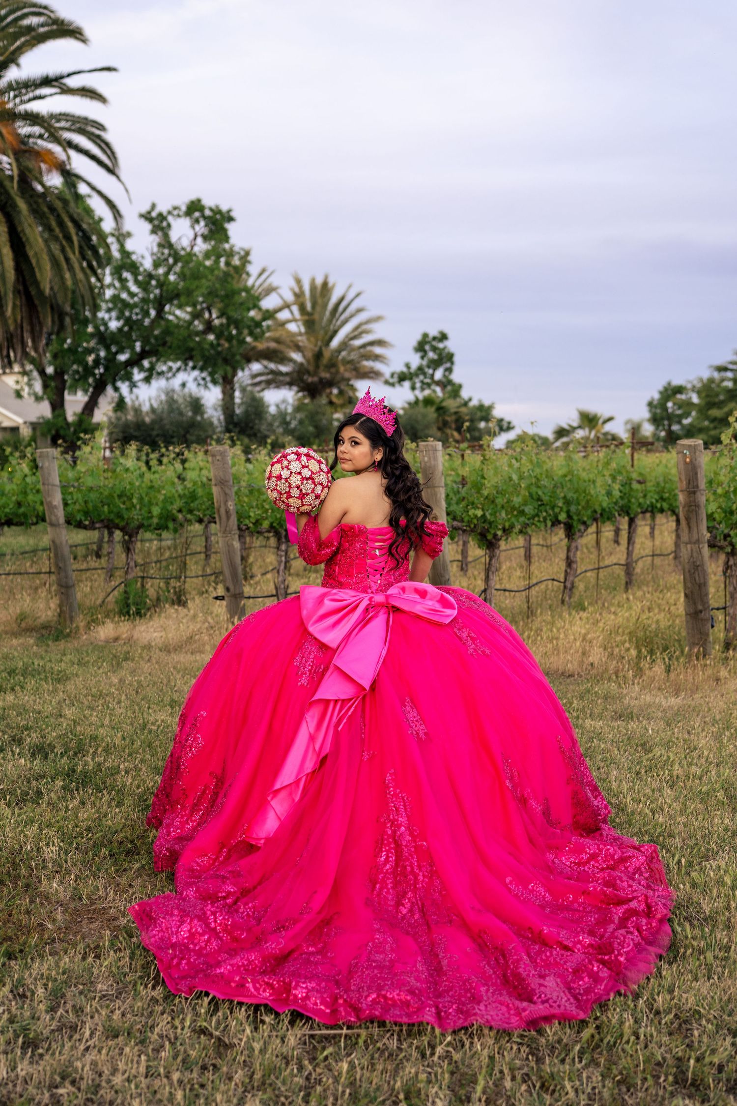 Quinceañera in hot pink ball gown with crown holds bouquet while posing in vineyard at dusk.