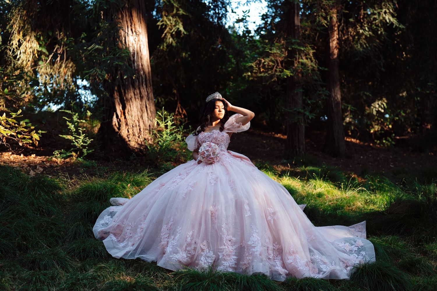 Woman in blush pink off-shoulder ball gown with floral details poses gracefully among tall redwood trees.
