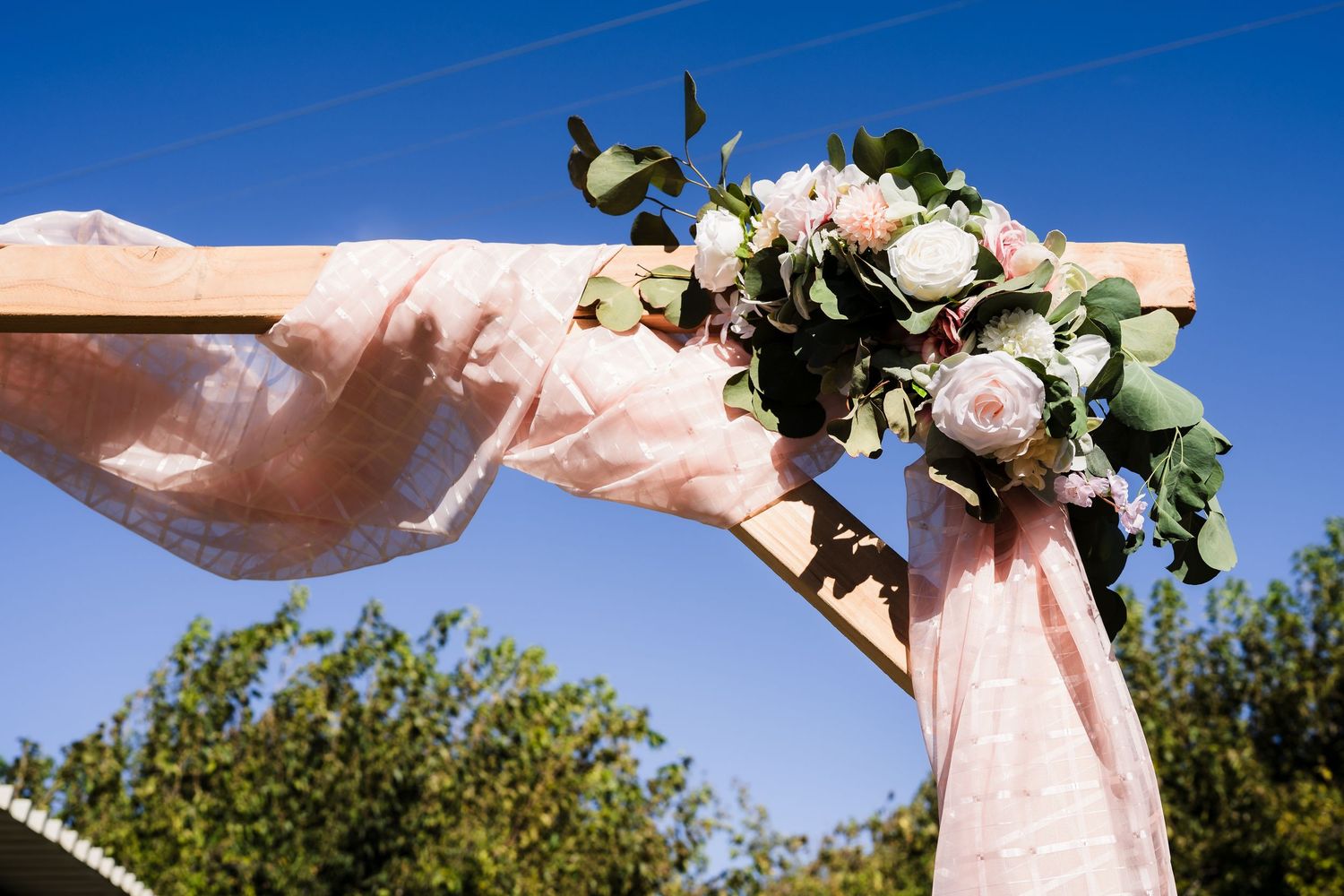 Wooden wedding arch adorned with pink fabric and blush rose bouquet against a clear blue sky.