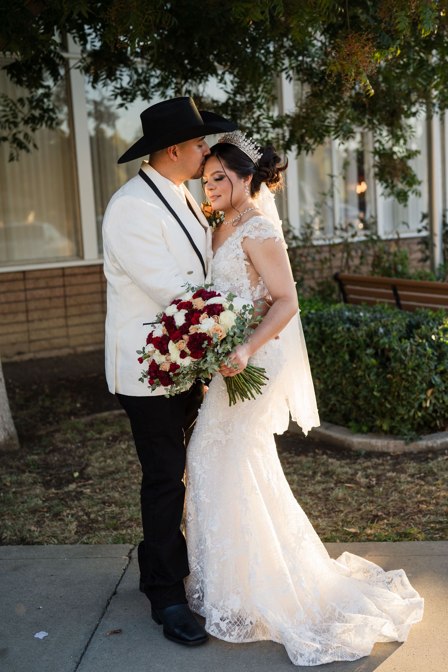 Bride in lace mermaid gown holding red roses bouquet, groom in white jacket and black cowboy hat, wedding portrait outdoors.