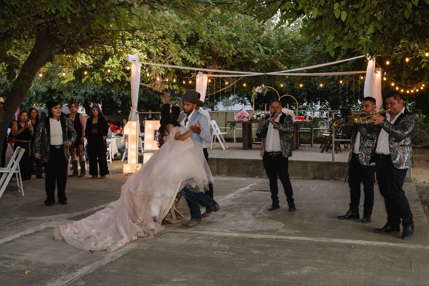 Bride and groom share first dance outdoors at evening wedding reception with live band and string lights.