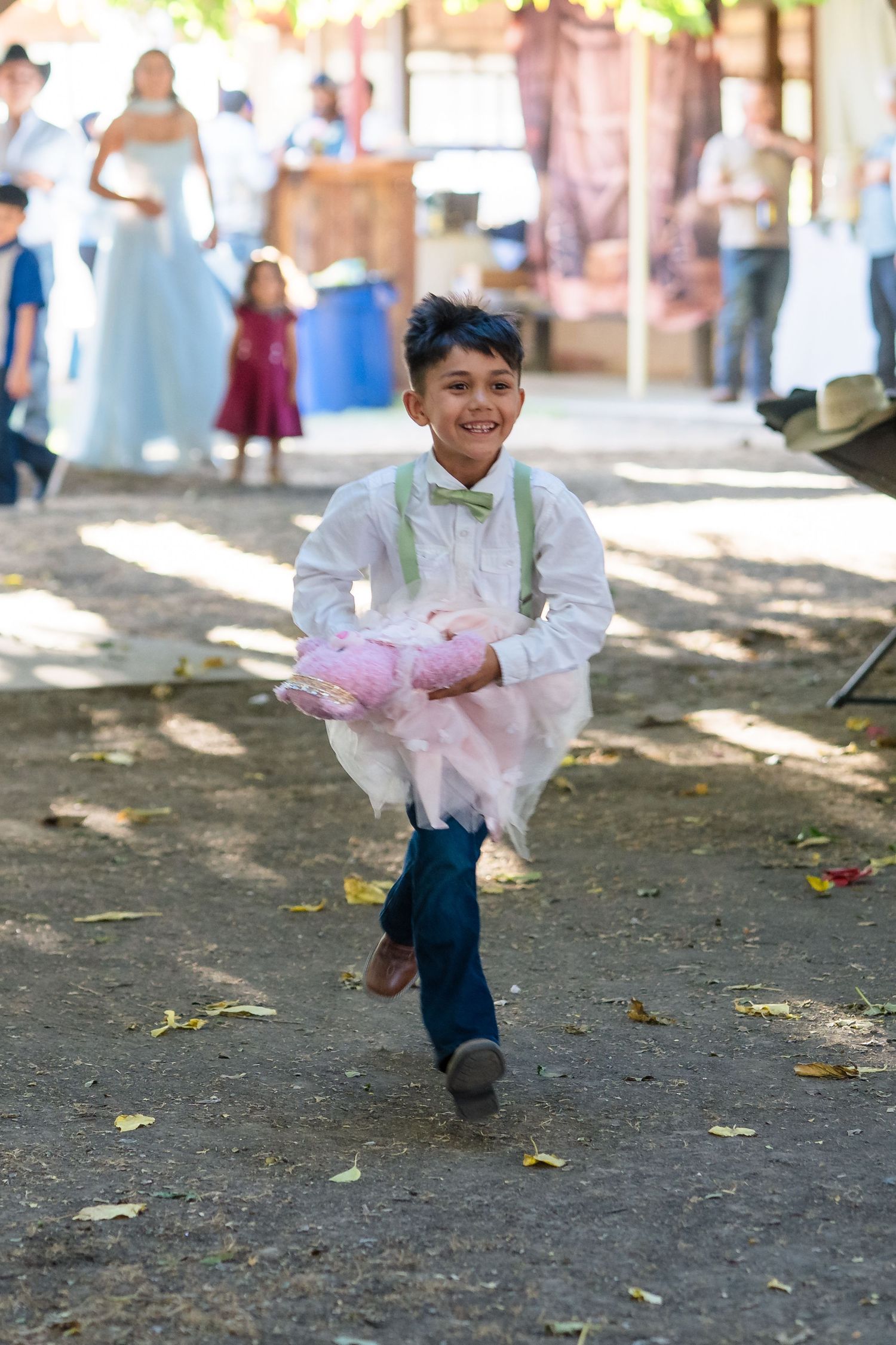 Smiling boy in white shirt, bow tie, and suspenders running at an outdoor wedding celebration.