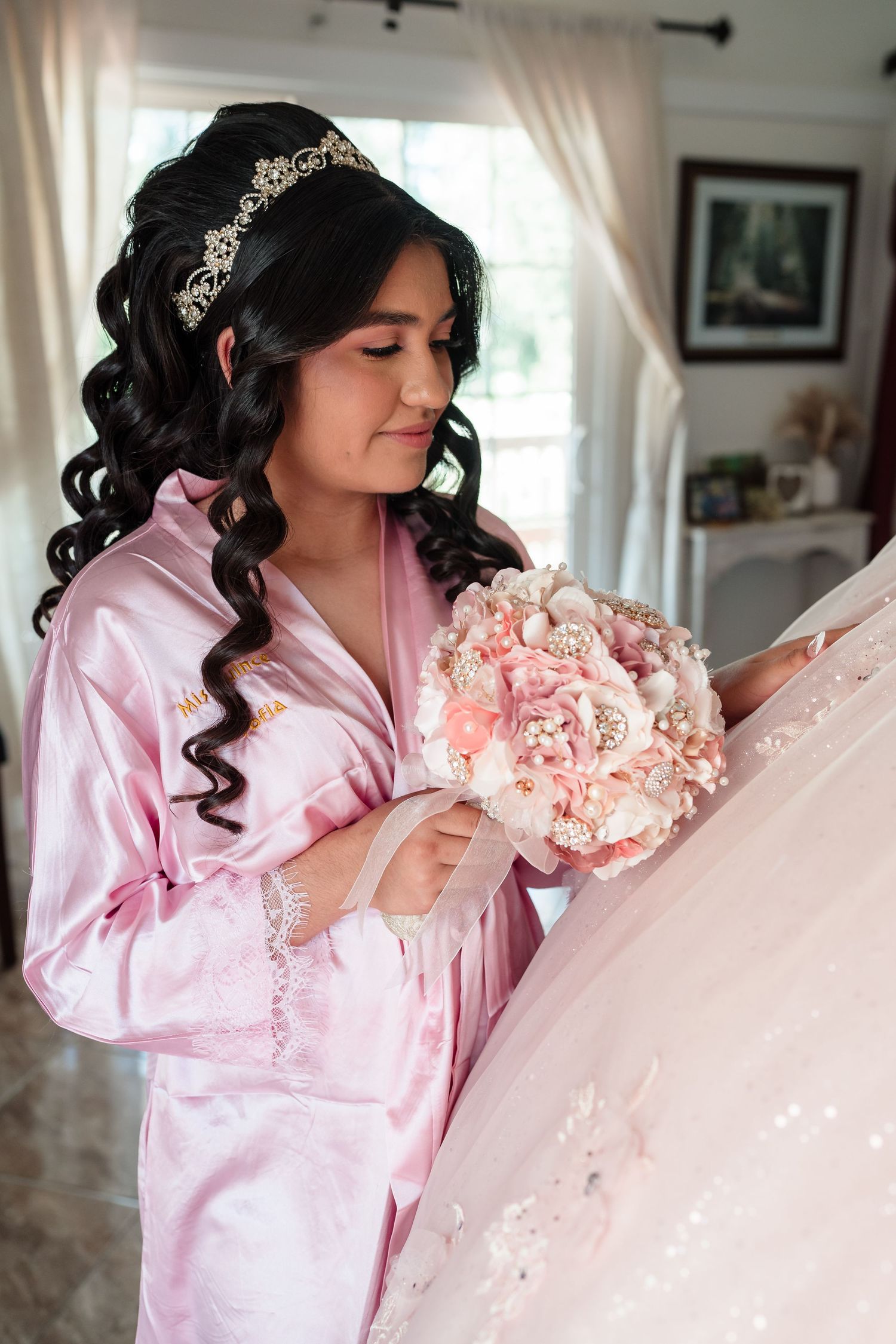 Young Latina in pink satin robe and tiara holds floral bouquet while admiring her quinceañera gown indoors.