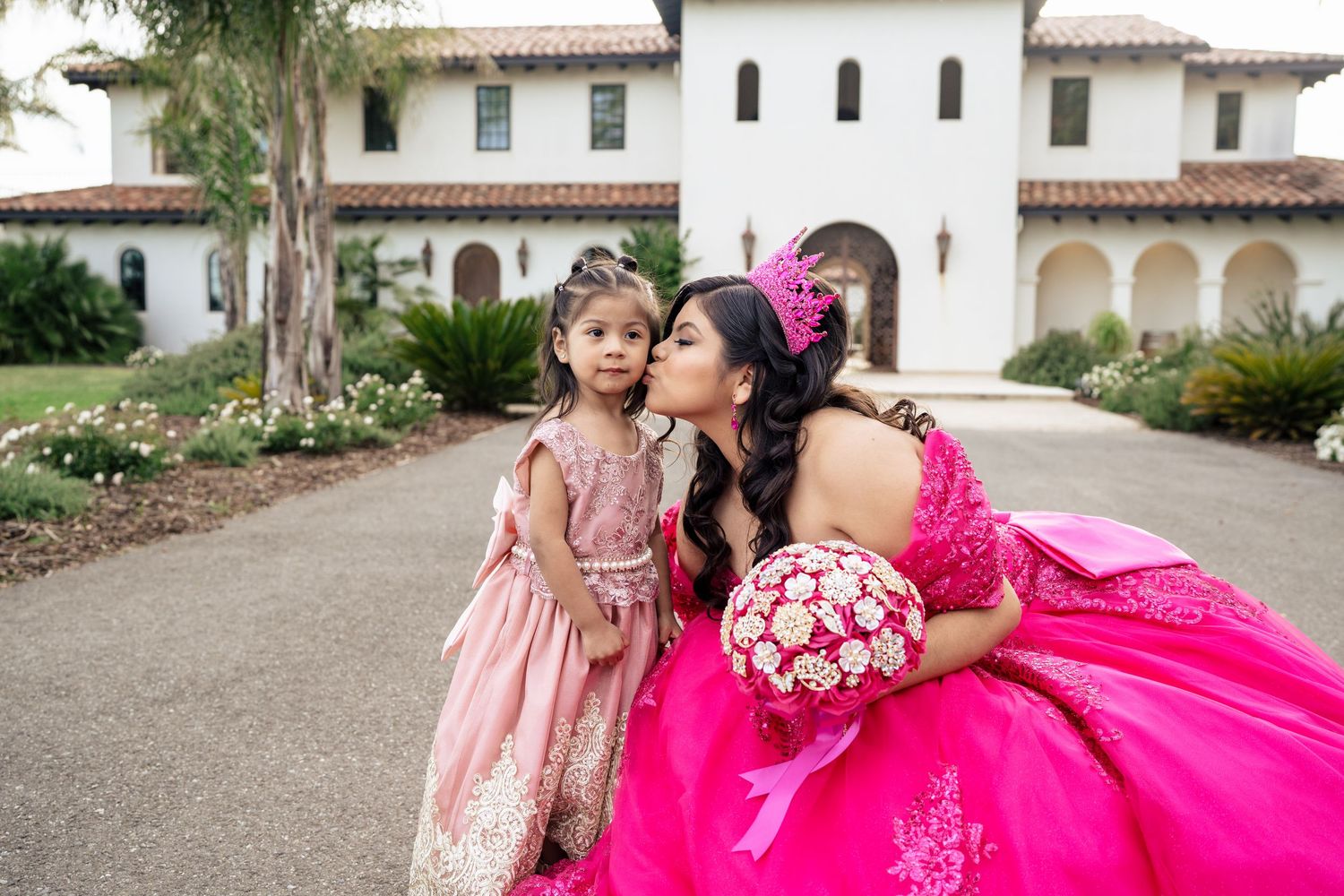 Quinceañera in hot pink gown kisses young girl in pink dress outside elegant white mansion.