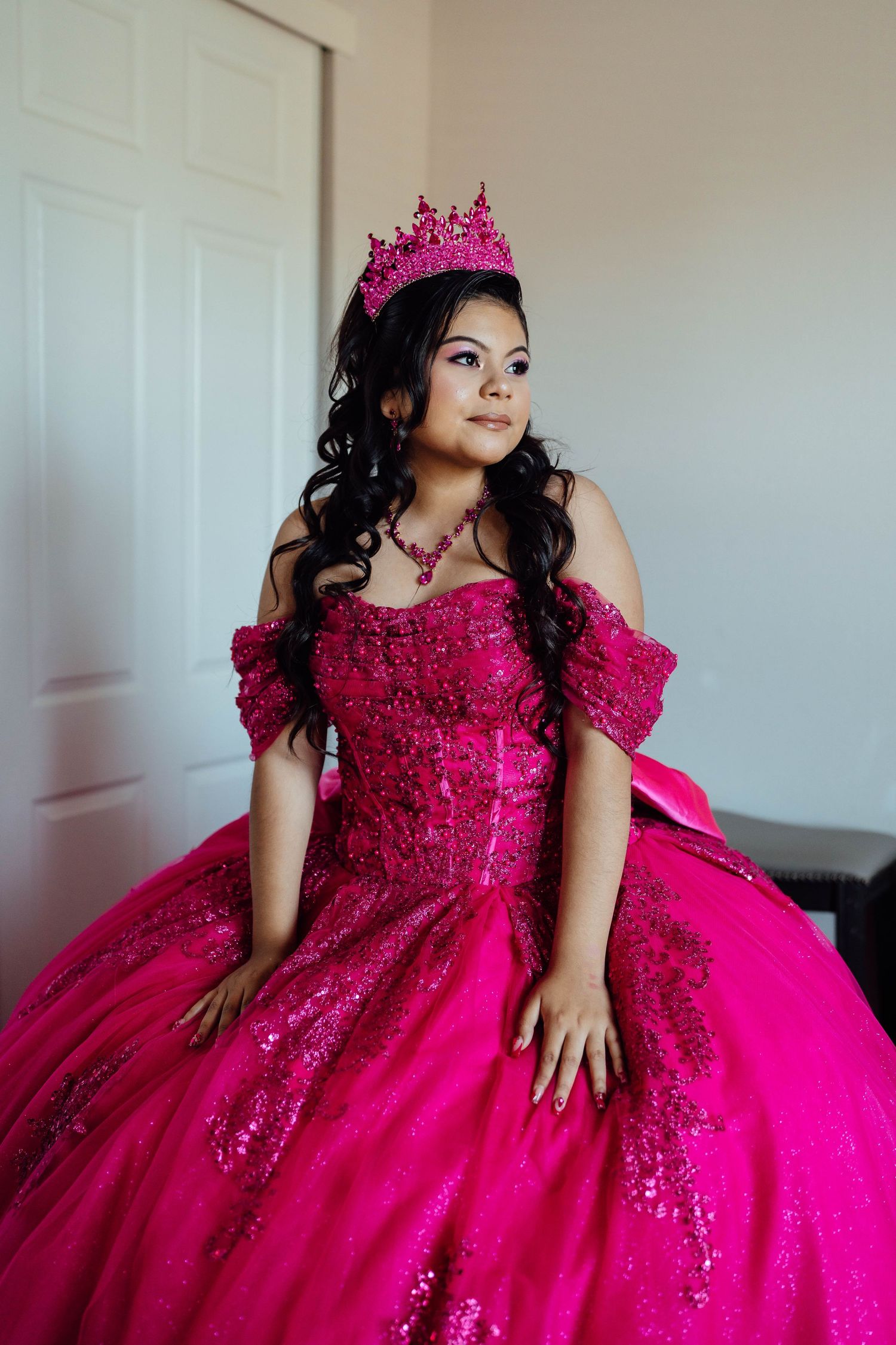 Young woman in sparkling hot pink quinceañera ball gown and crown posing indoors.