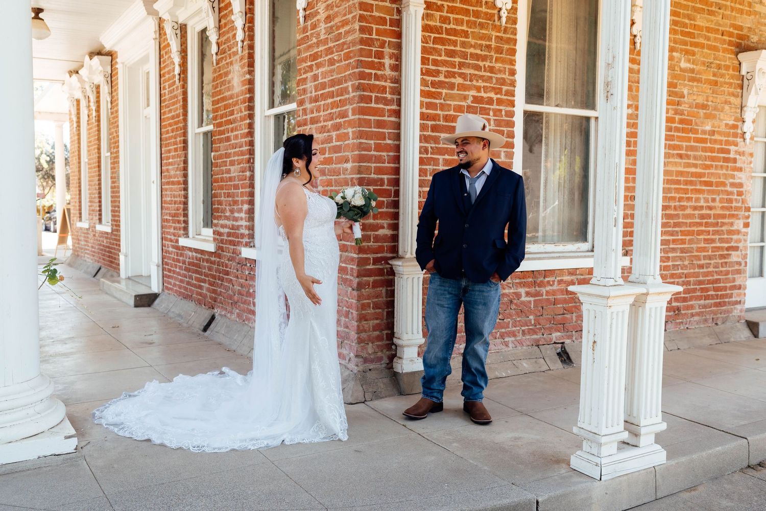 Bride in a white gown and groom in a cowboy hat hold hands outside a historic brick building.