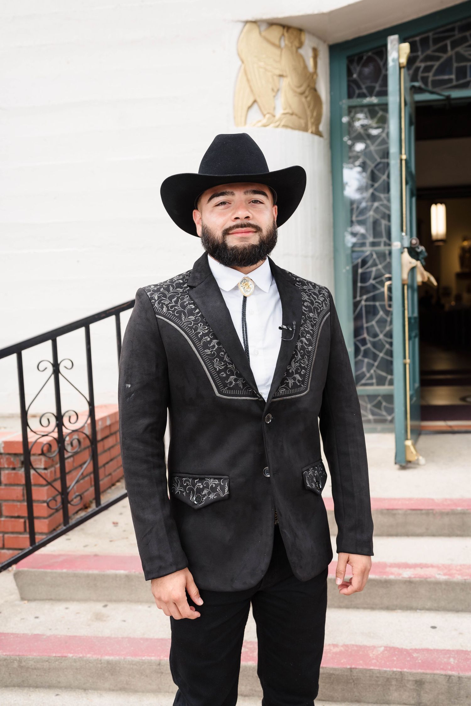 Bearded man in black cowboy hat and embroidered western suit jacket posing outside a building.
