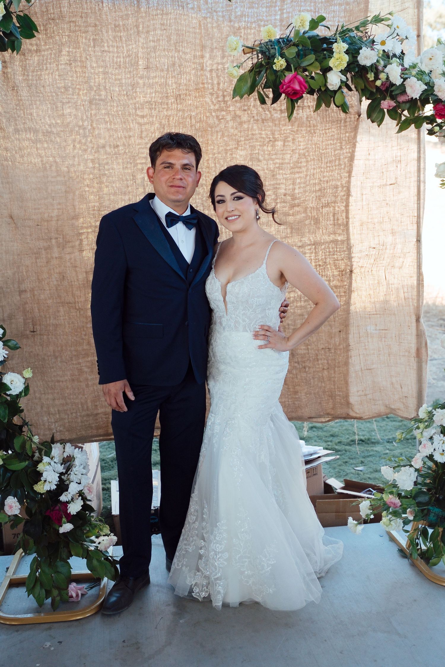Bride in white lace gown and groom in black tuxedo pose together at floral wedding altar with burlap backdrop.