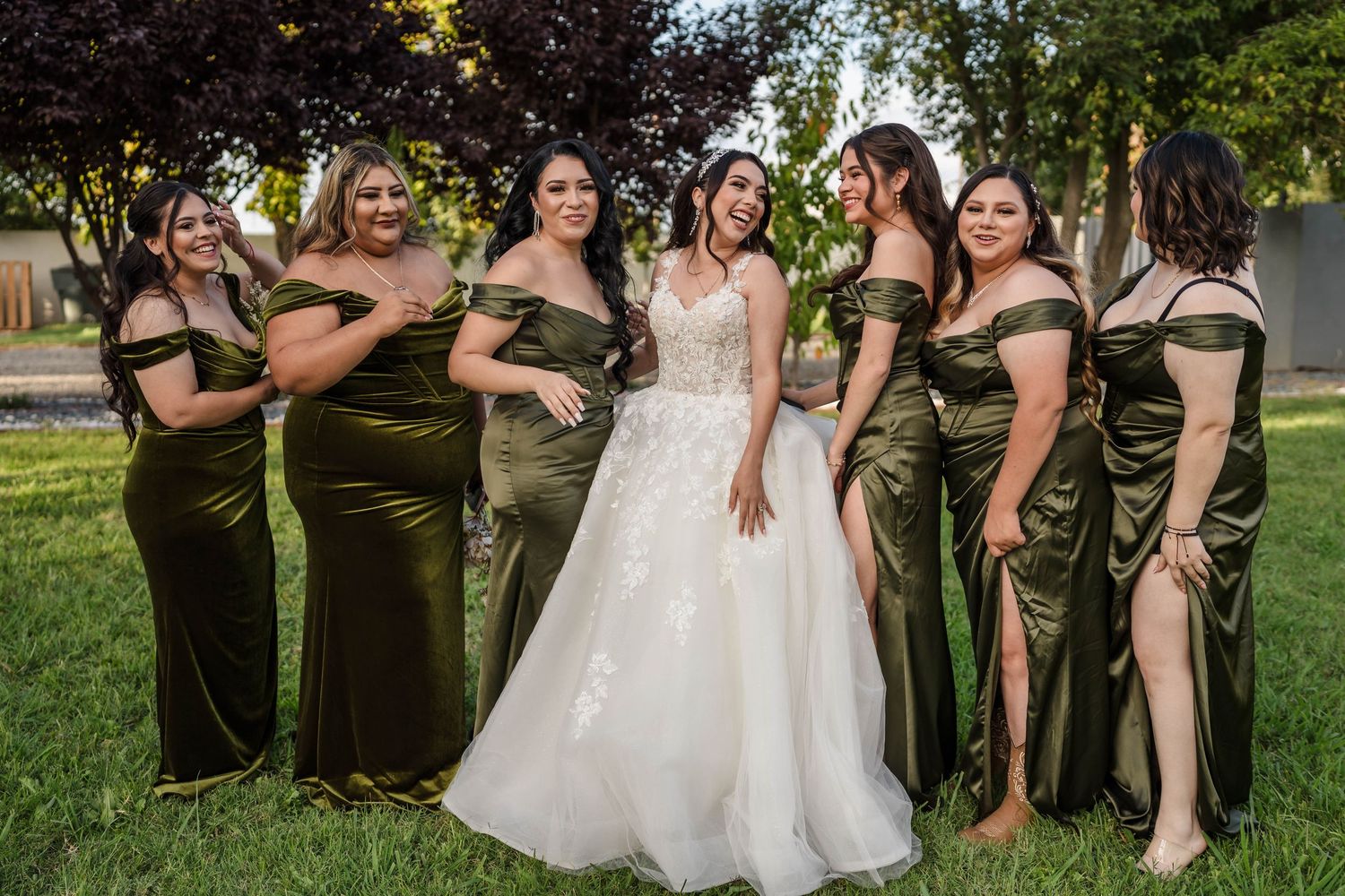 Bride in white lace gown poses with six bridesmaids in olive green satin dresses on a sunny lawn.