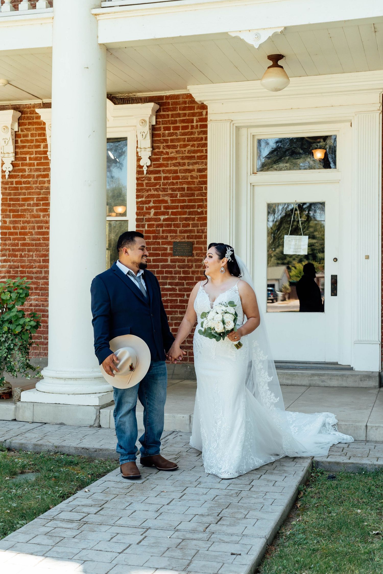 Bride and groom hold hands walking along brick path in front of a historic white-columned mansion.