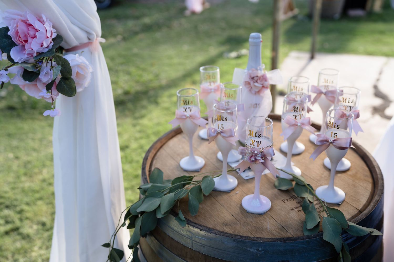 Elegant wedding champagne flutes with pink floral decorations displayed on a rustic wooden barrel outdoors.