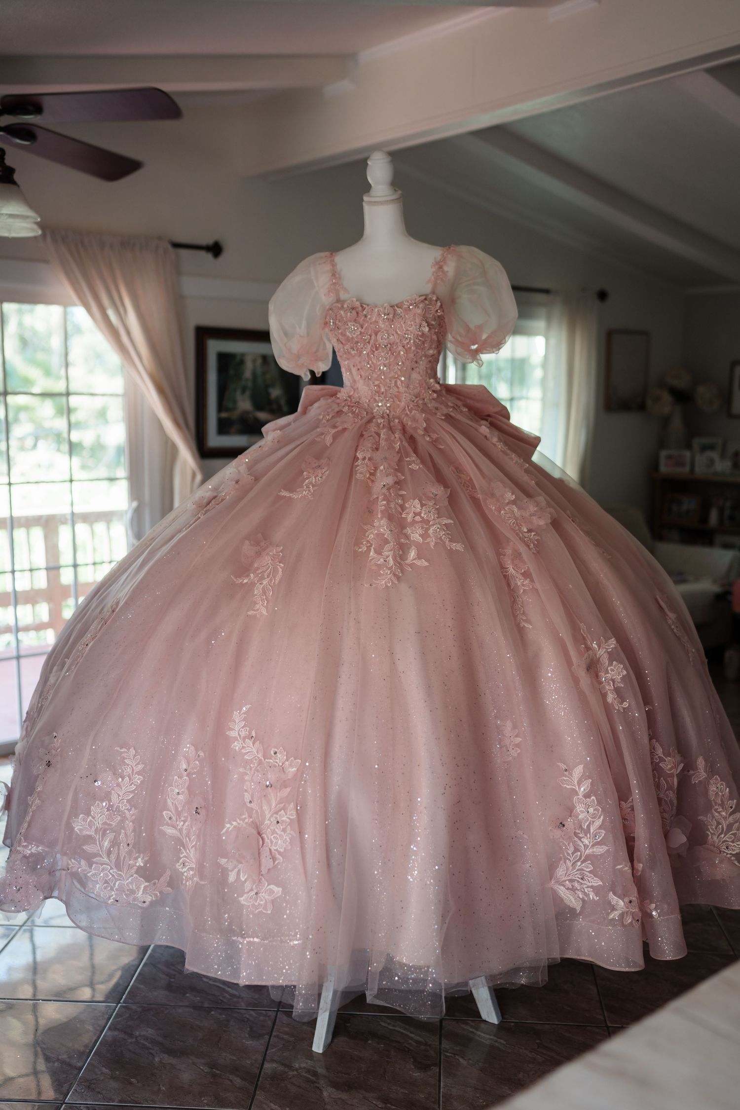 Elegant blush pink ball gown with lace appliques and puff sleeves displayed on a mannequin indoors.