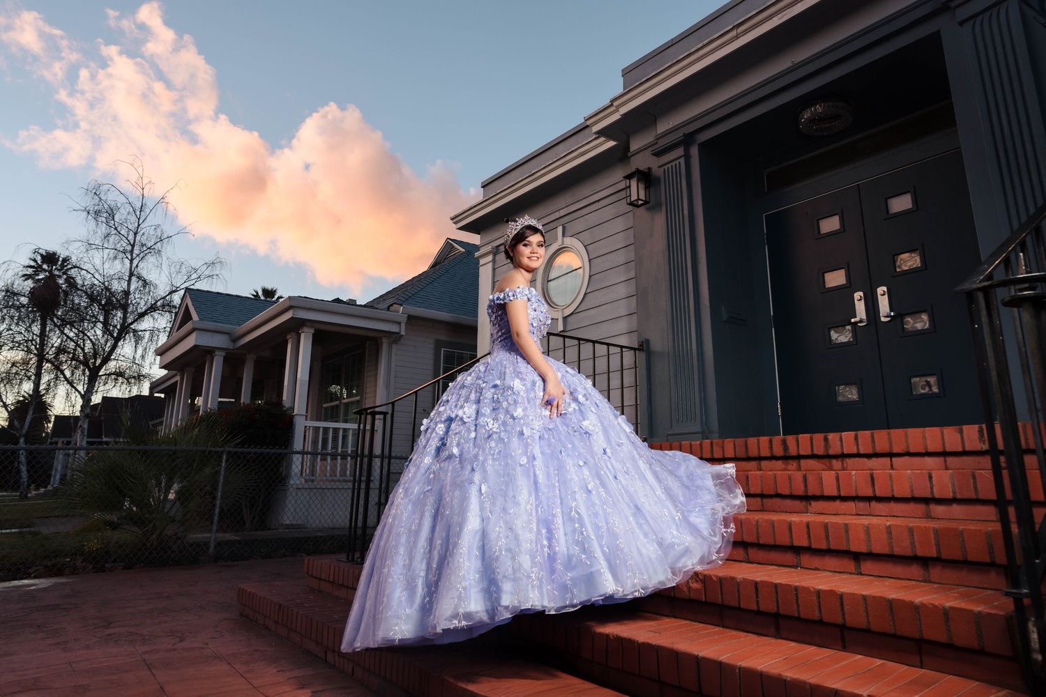 Young woman in a lavender quinceañera ball gown posing on brick steps of a house at sunset.