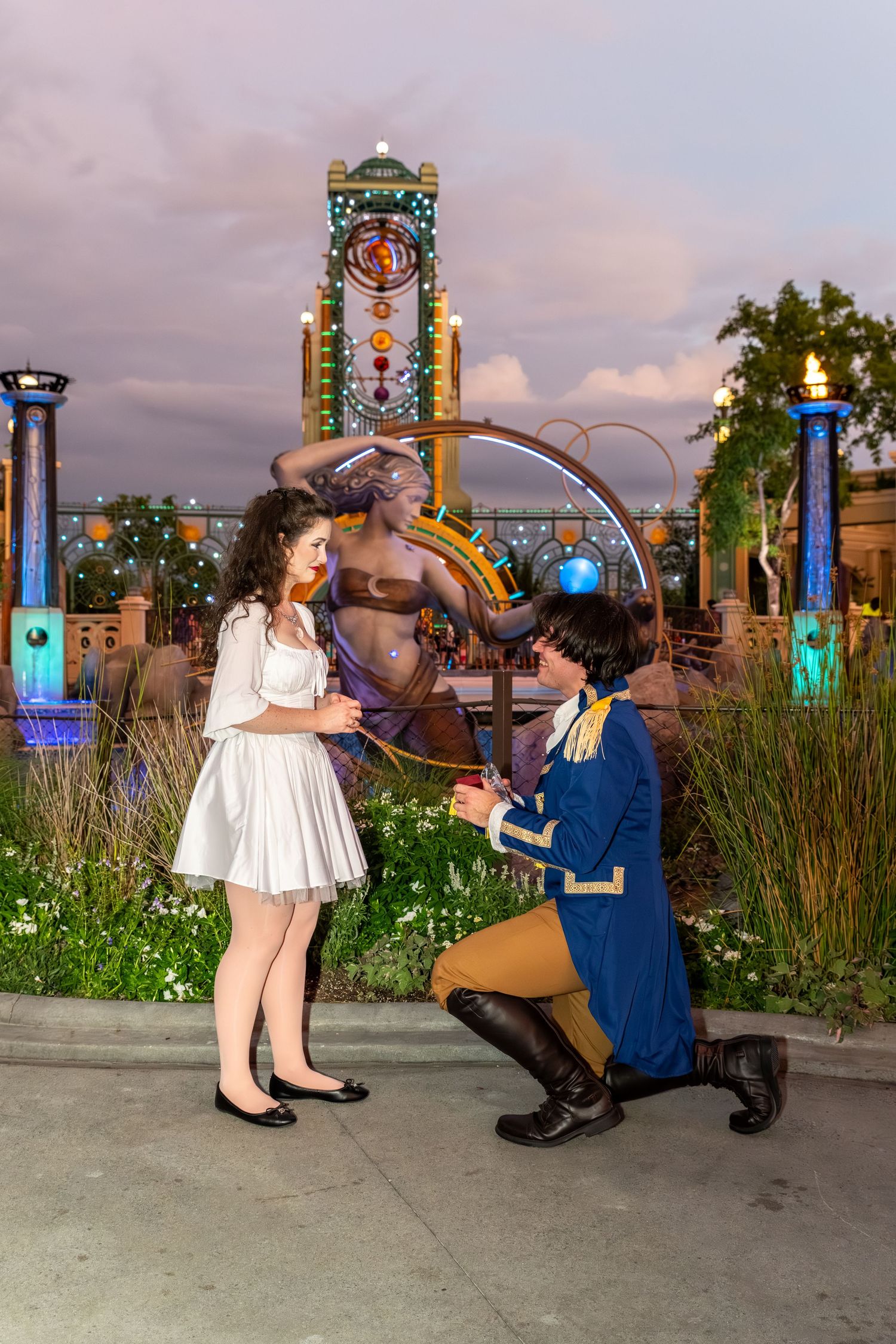 Man on one knee proposing to woman in white dress at a Disney theme park at dusk.