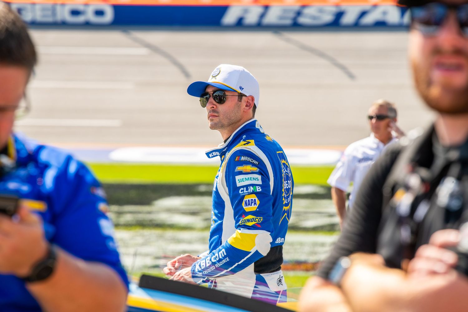 NASCAR driver in NAPA Auto Parts racing suit walks pit road at a sunny speedway during a race event.
