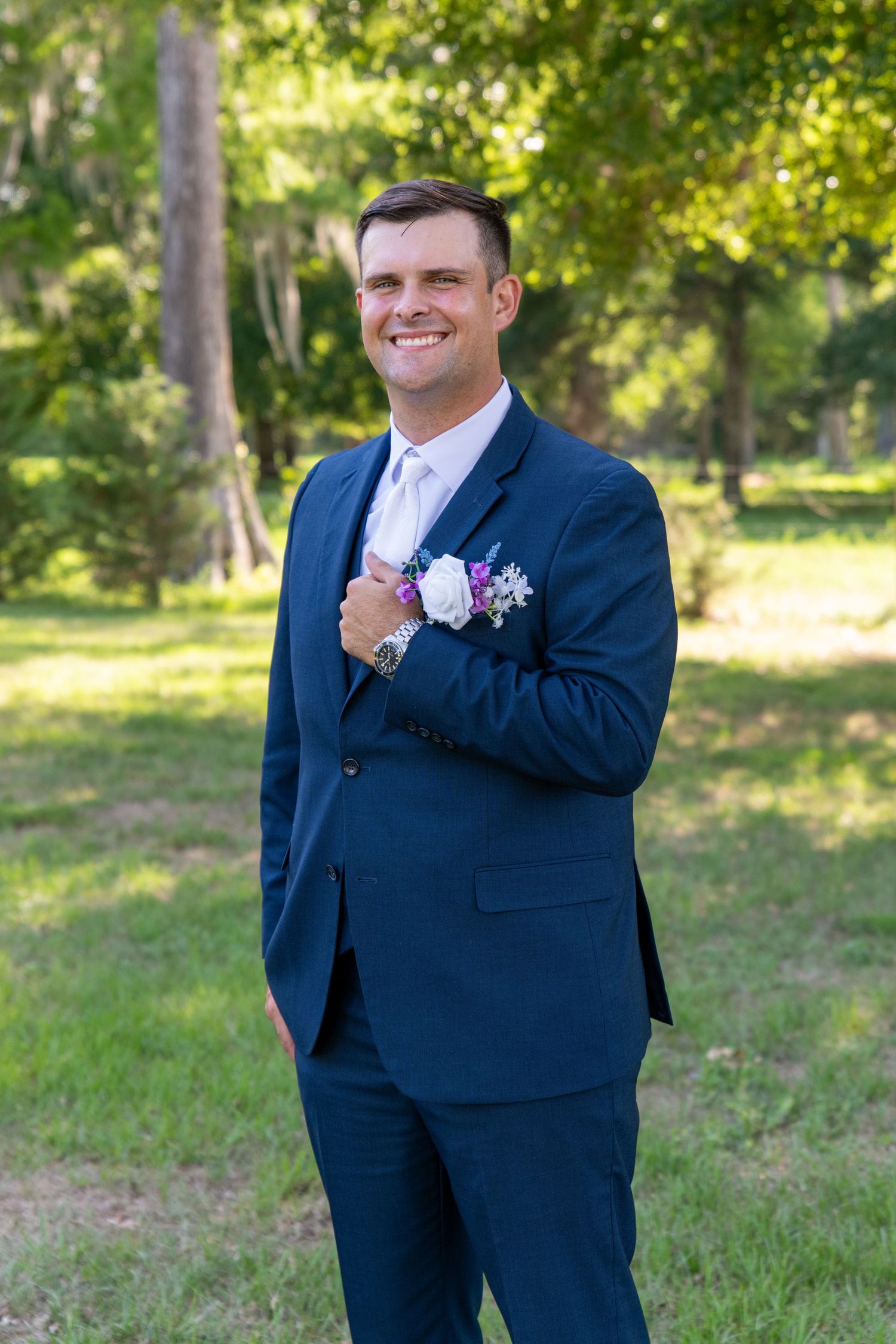 Smiling groom in navy blue suit with floral boutonniere standing outdoors in a lush green park setting.