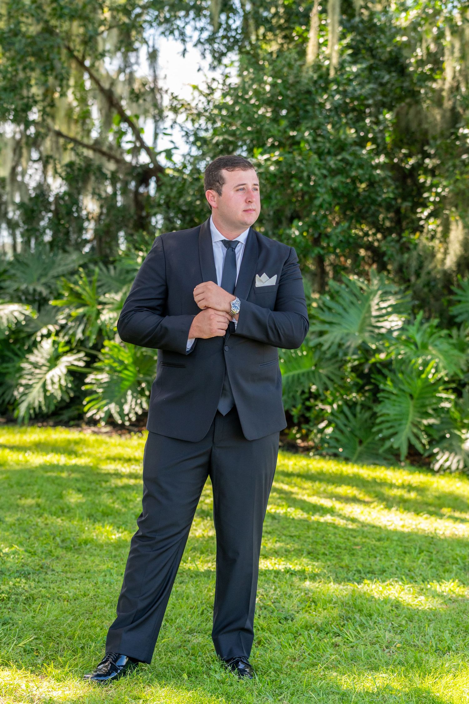 A groom in a dark suit adjusts his cufflinks outdoors on green grass with lush tropical trees in the background.