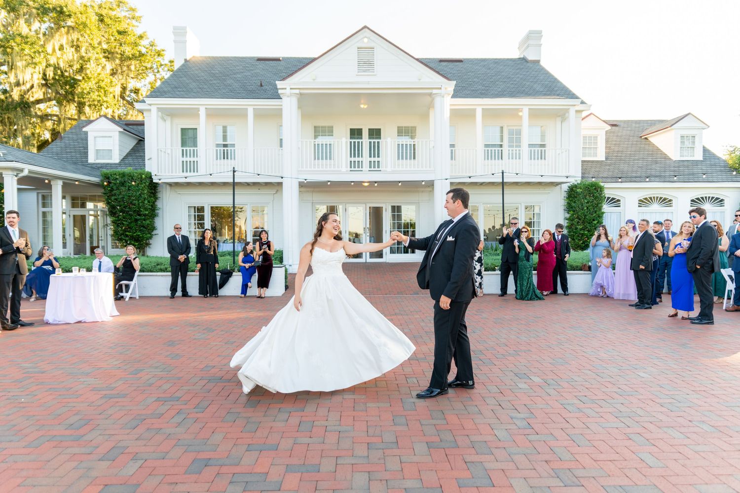 Bride and groom share first dance on brick patio outside elegant white colonial wedding venue with guests watching.