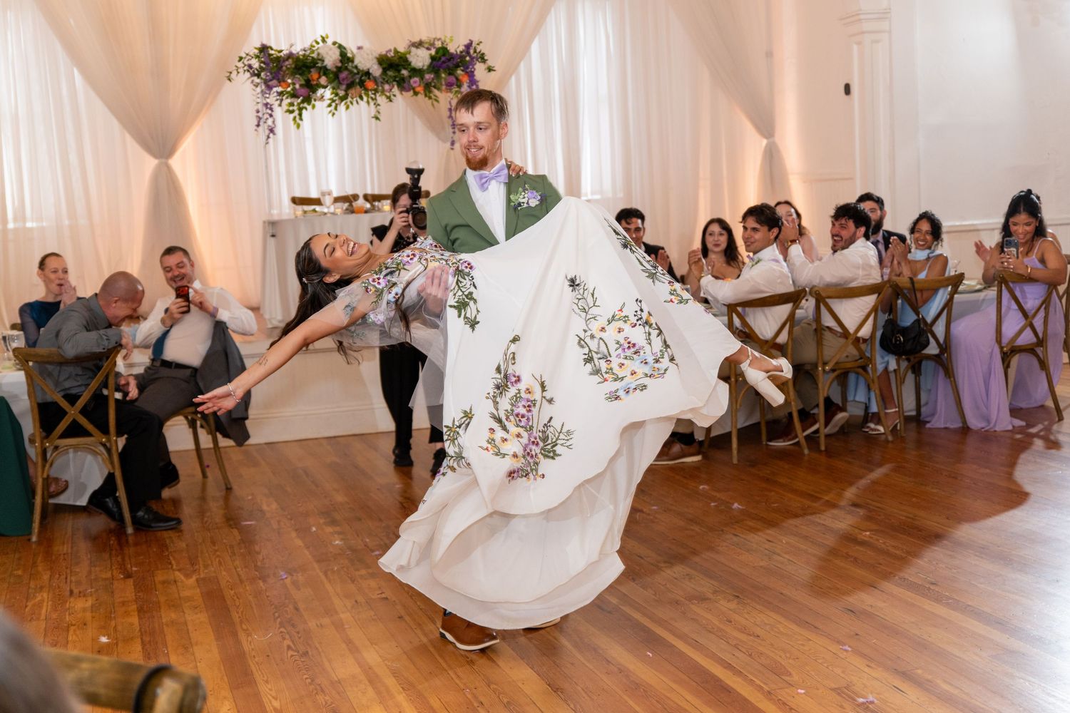 Groom twirling bride in floral wedding gown on wooden dance floor during their wedding reception first dance.