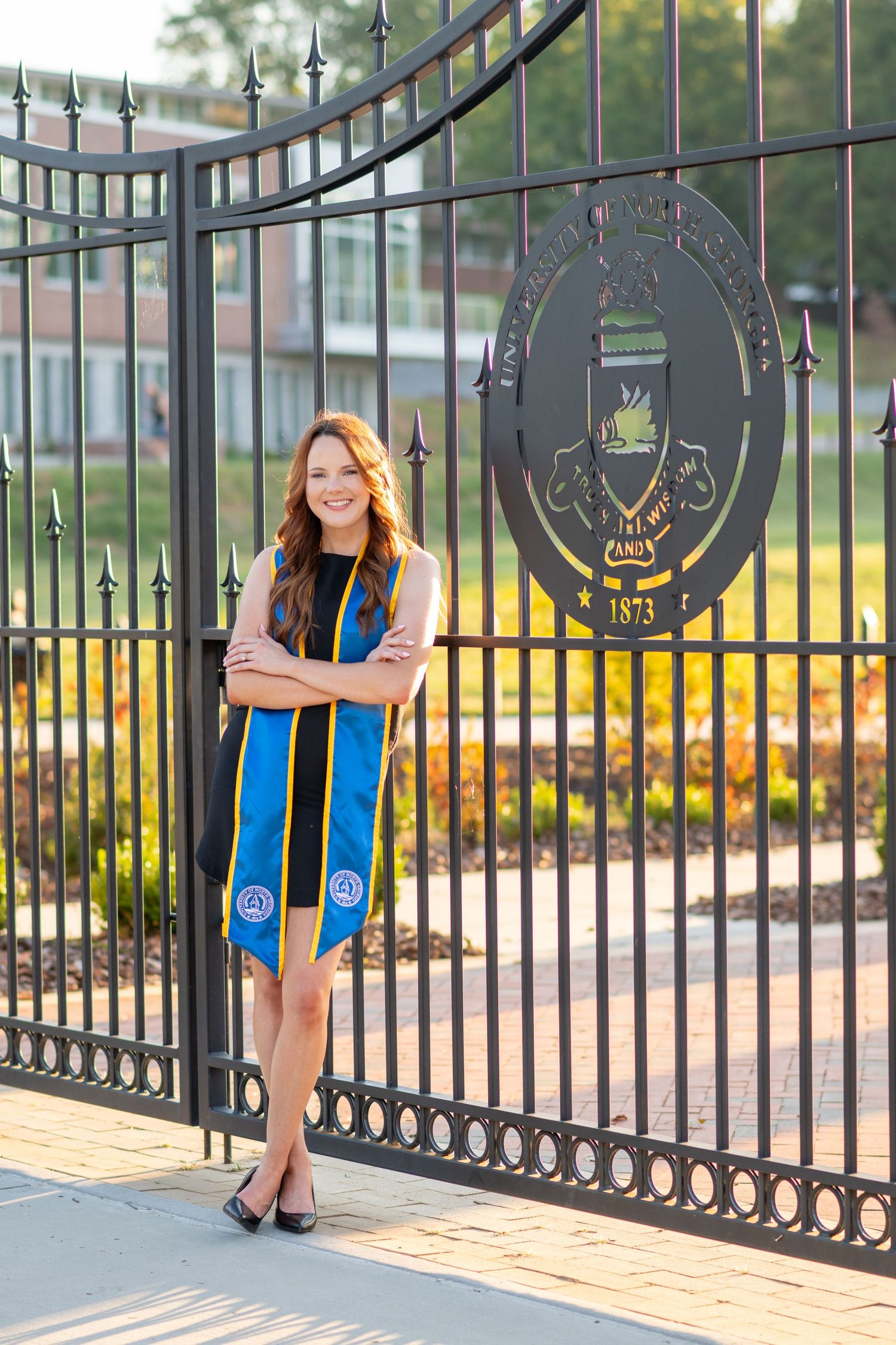 Smiling graduate poses in blue and gold stole before university gate with school seal dated 1873.