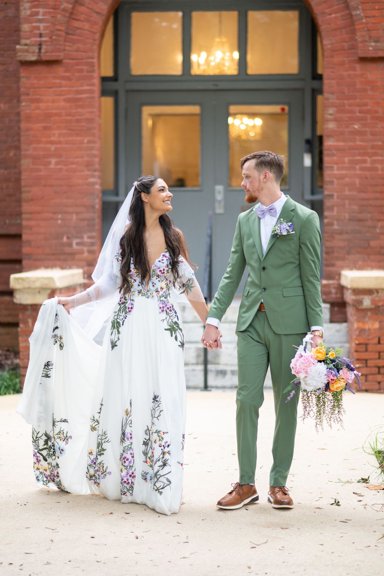 Bride in floral white gown and groom in green suit hold hands outside a brick building on their wedding day.