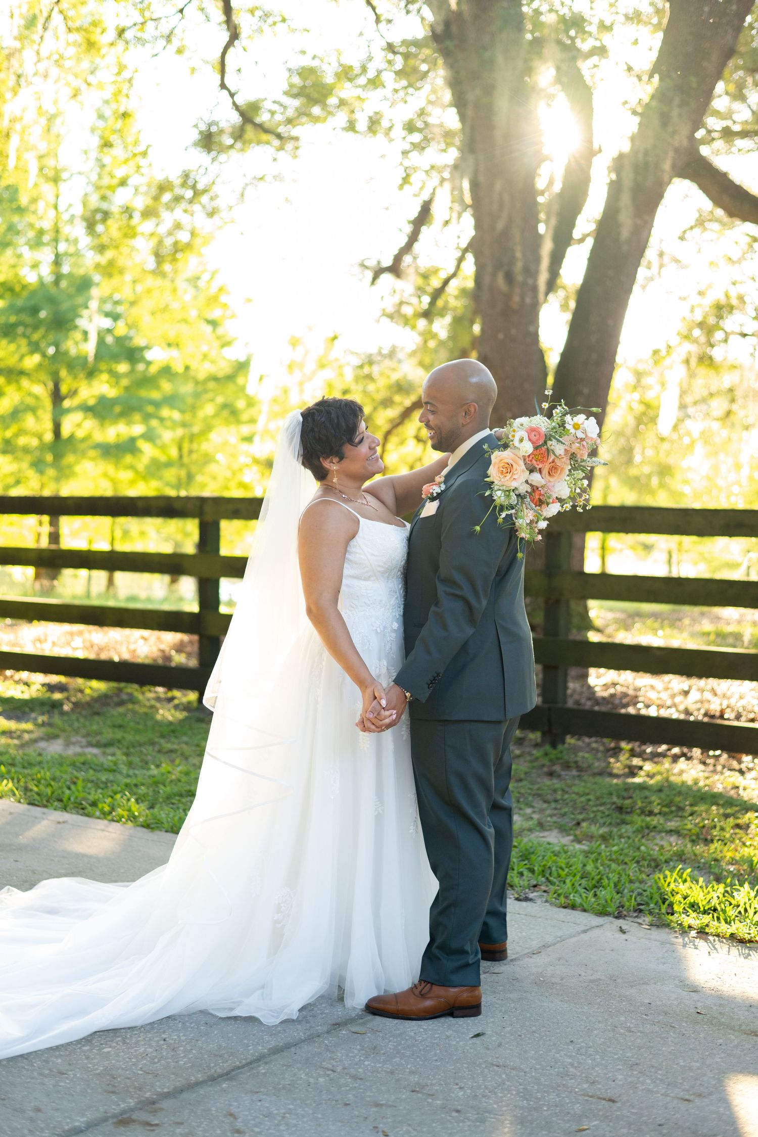 Bride and groom holding hands outdoors, bride in white gown with veil, groom in gray suit, golden sunlight through trees.