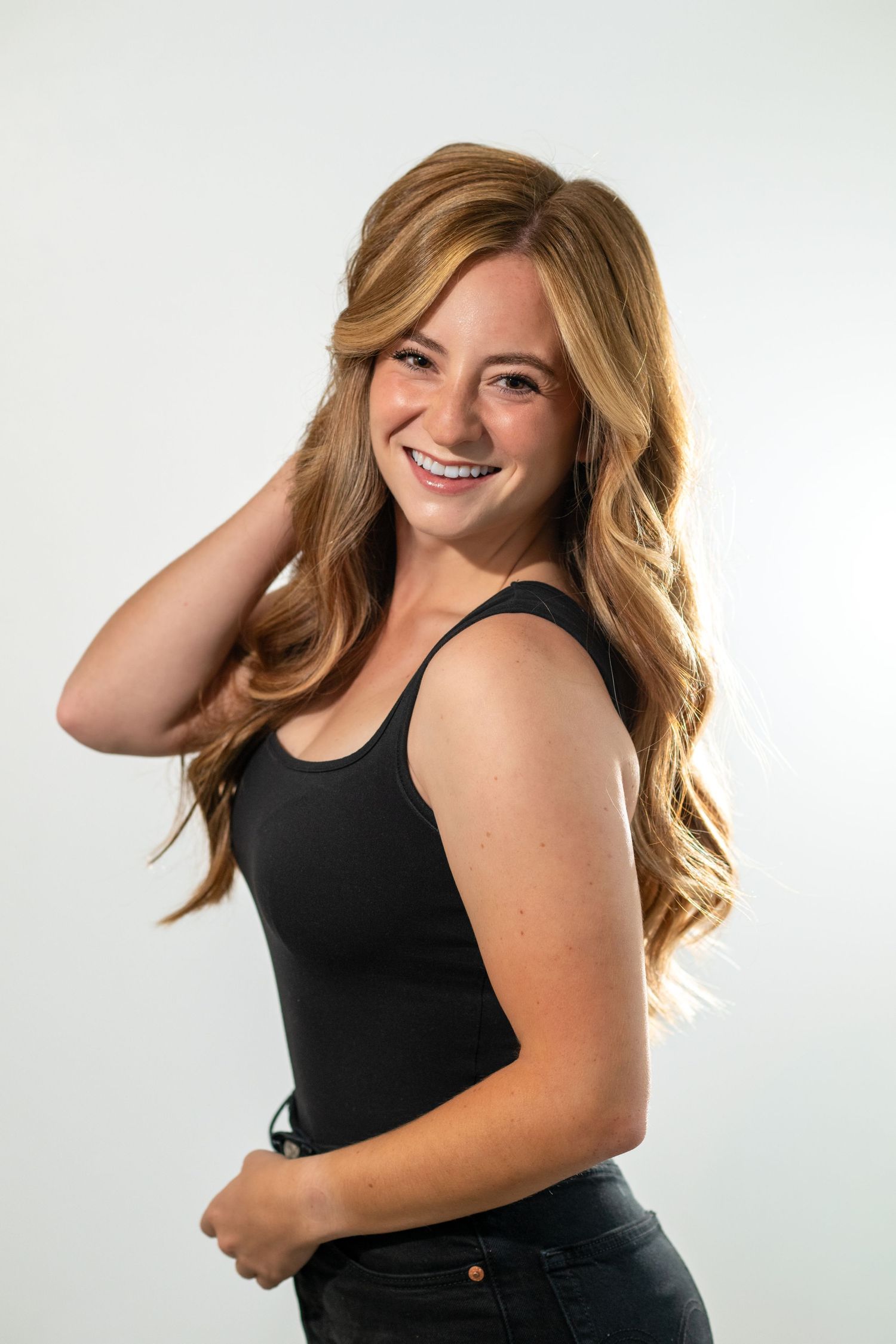 Smiling young woman with long wavy blonde hair wearing a black tank top posing against a light gray background.
