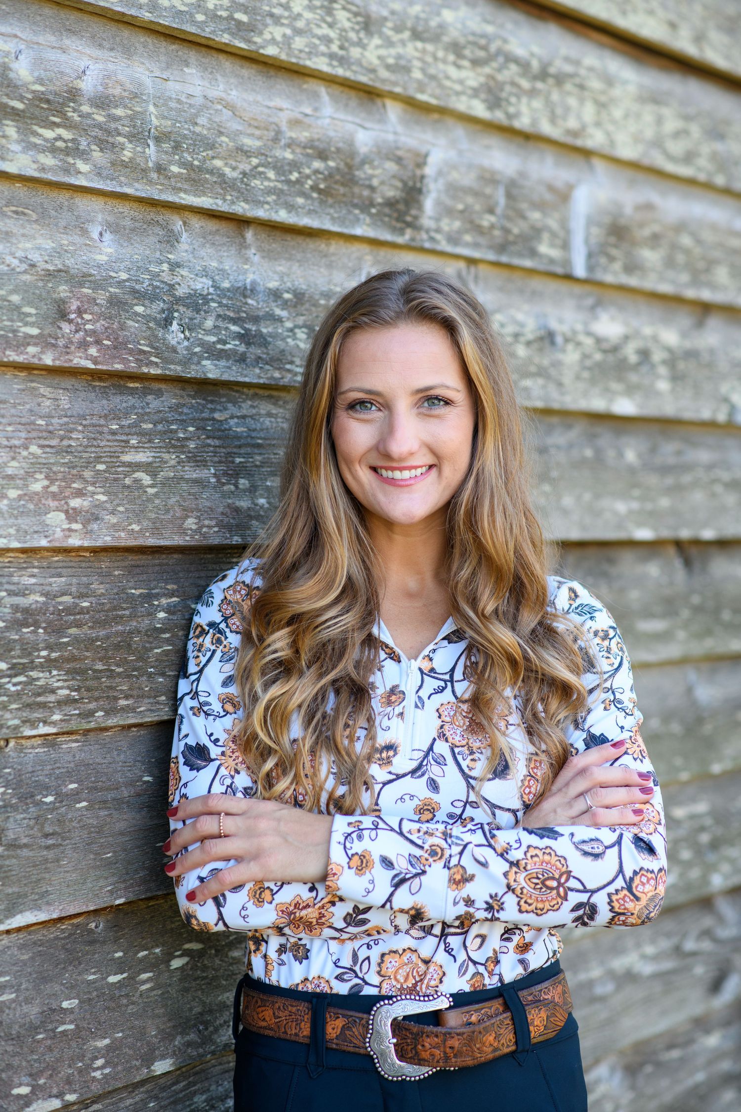Smiling young woman with long wavy hair in floral shirt, arms crossed, leaning against rustic wooden wall.