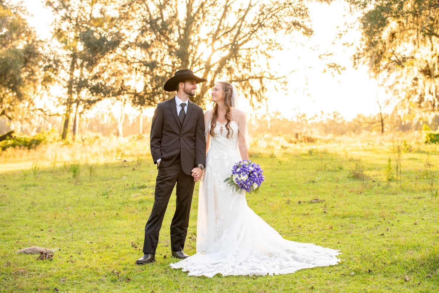 Bride and groom holding hands in a sunlit field, she holds purple bouquet in white gown, he wears dark suit and cowboy hat.