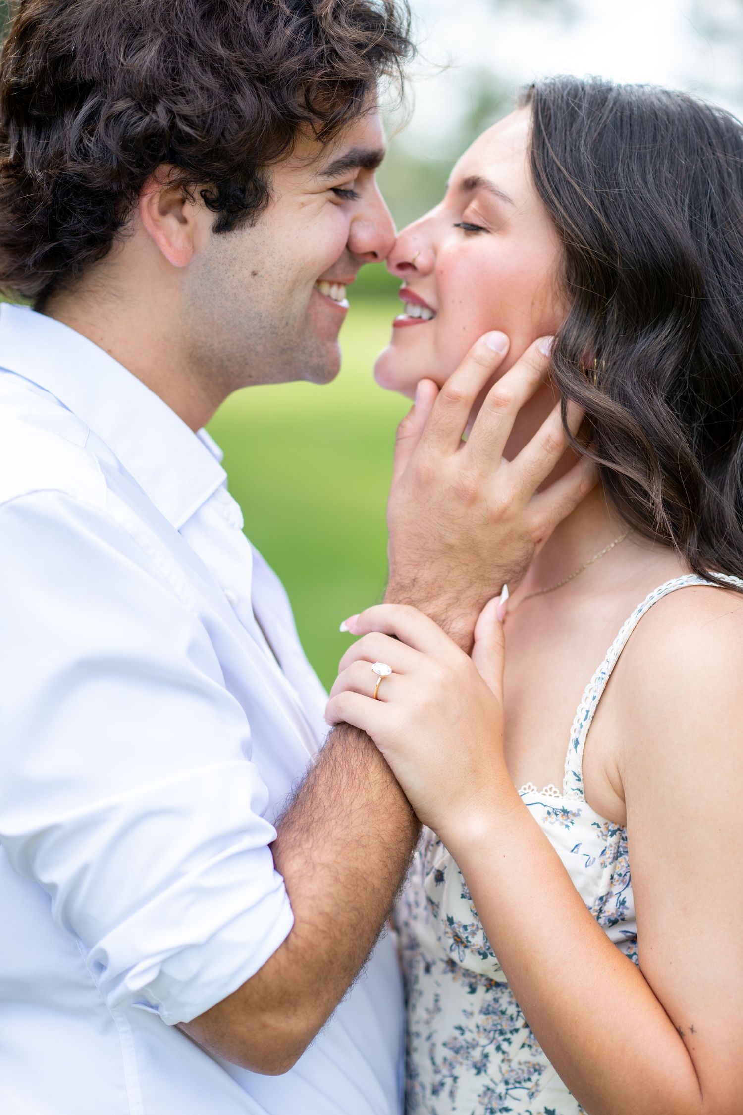 Joyful couple about to kiss outdoors, woman wearing diamond engagement ring touching man's face.