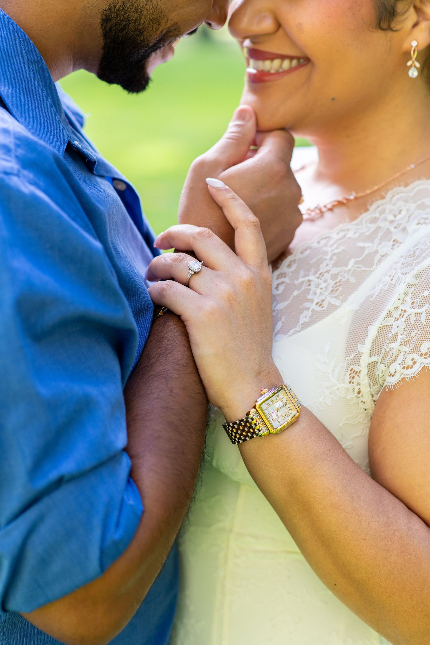 Couple holding hands closeup showing engagement ring and gold watch, woman in lace dress smiling at man in blue shirt.
