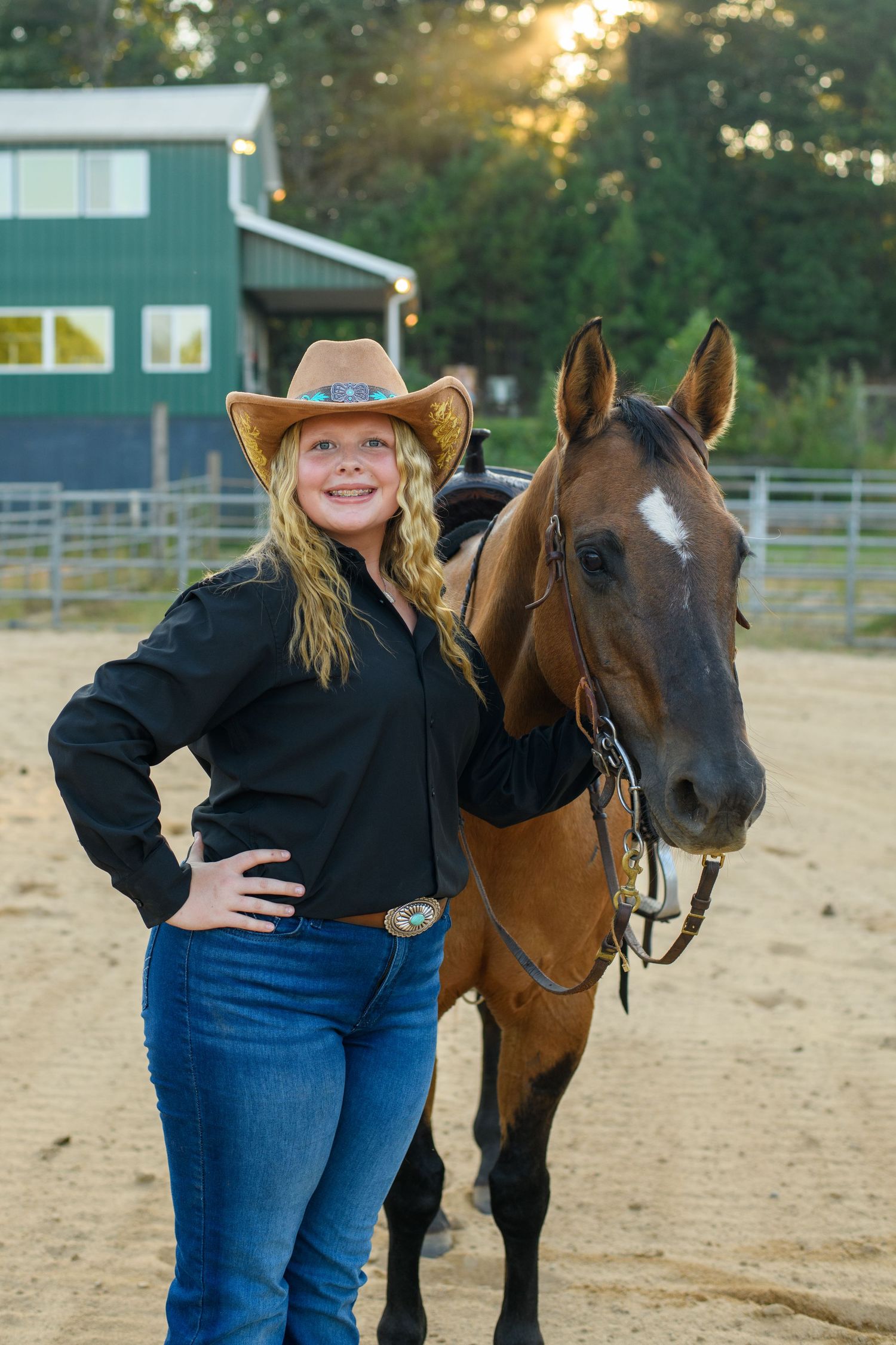 Smiling blonde woman in cowboy hat and black shirt posing with a bay horse at a sandy ranch arena.