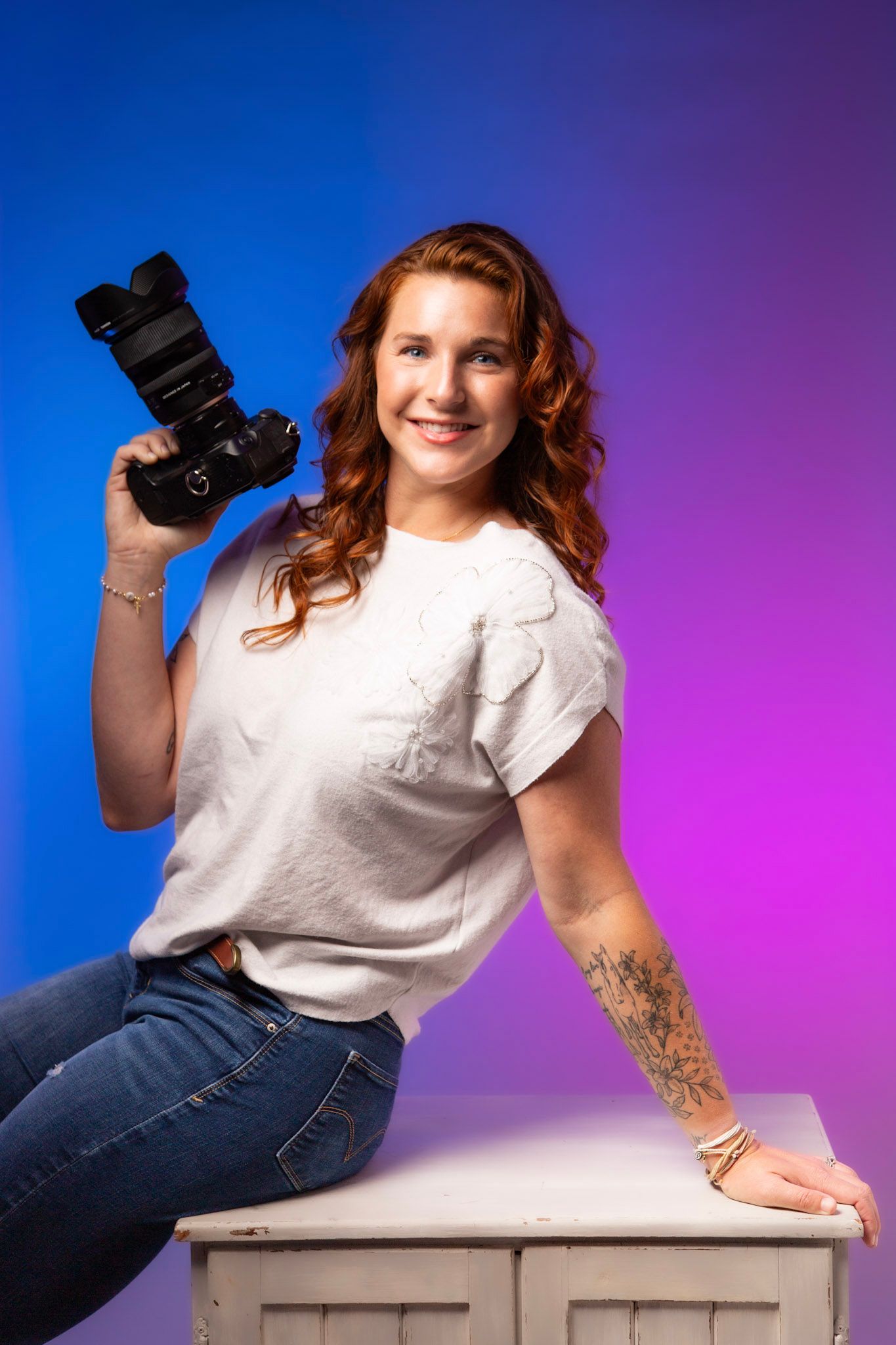 Smiling redhead woman holding a DSLR camera, sitting on white furniture with a colorful blue and pink studio background.