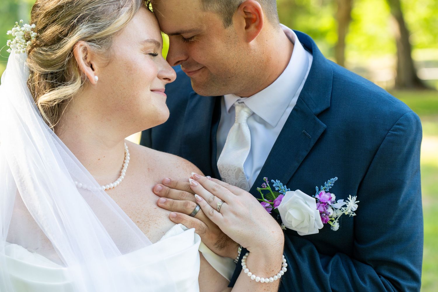 Bride and groom sharing an intimate moment, she wears pearl jewelry and veil, he wears navy suit with floral boutonniere.