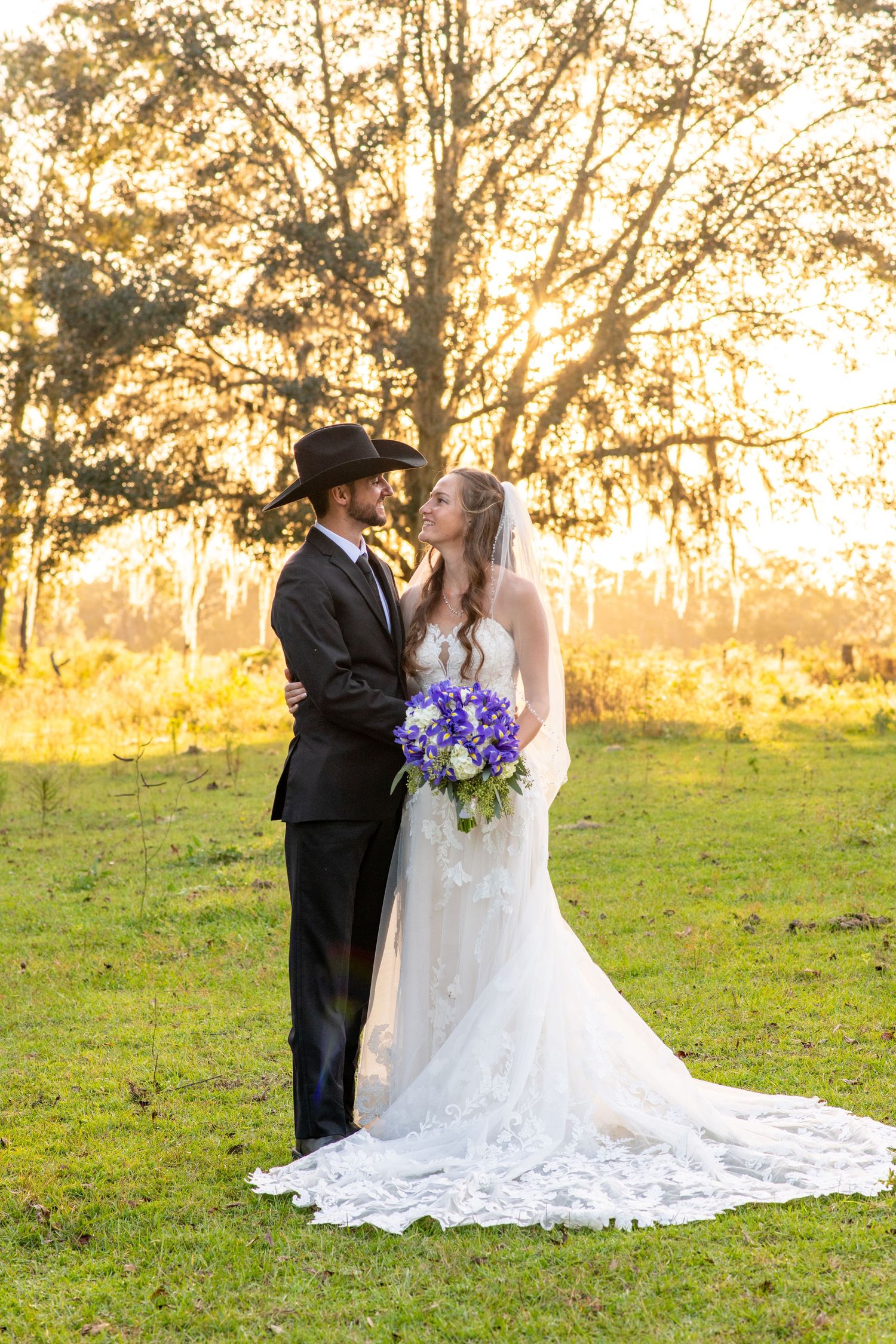 Bride and groom share a romantic sunset moment outdoors, she holds purple bouquet in lace gown, he wears black suit and cowboy hat.