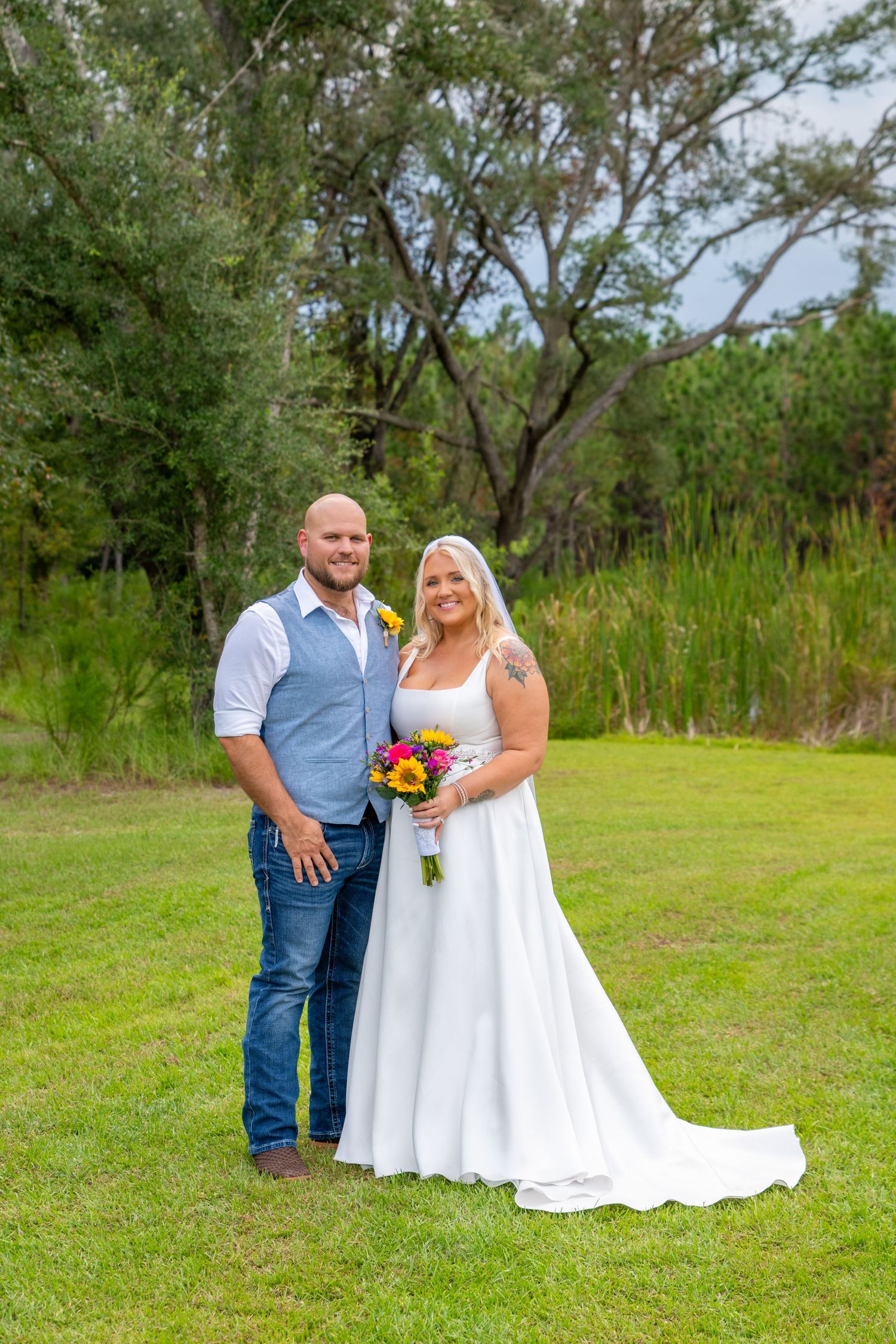 Bride in white gown holding colorful bouquet poses with groom in blue vest outdoors.