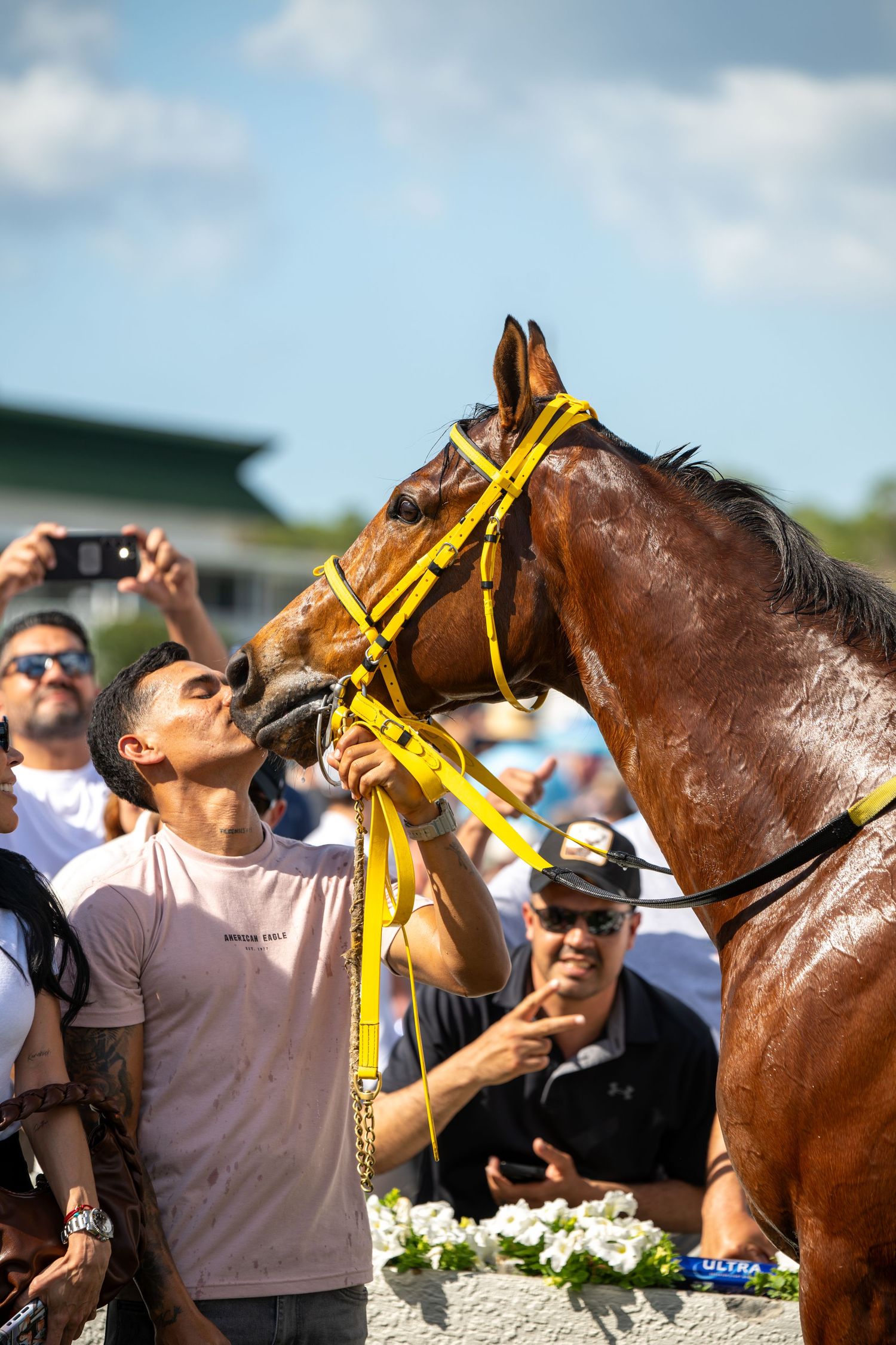 Man kisses bay thoroughbred horse wearing yellow halter surrounded by crowd at racetrack.