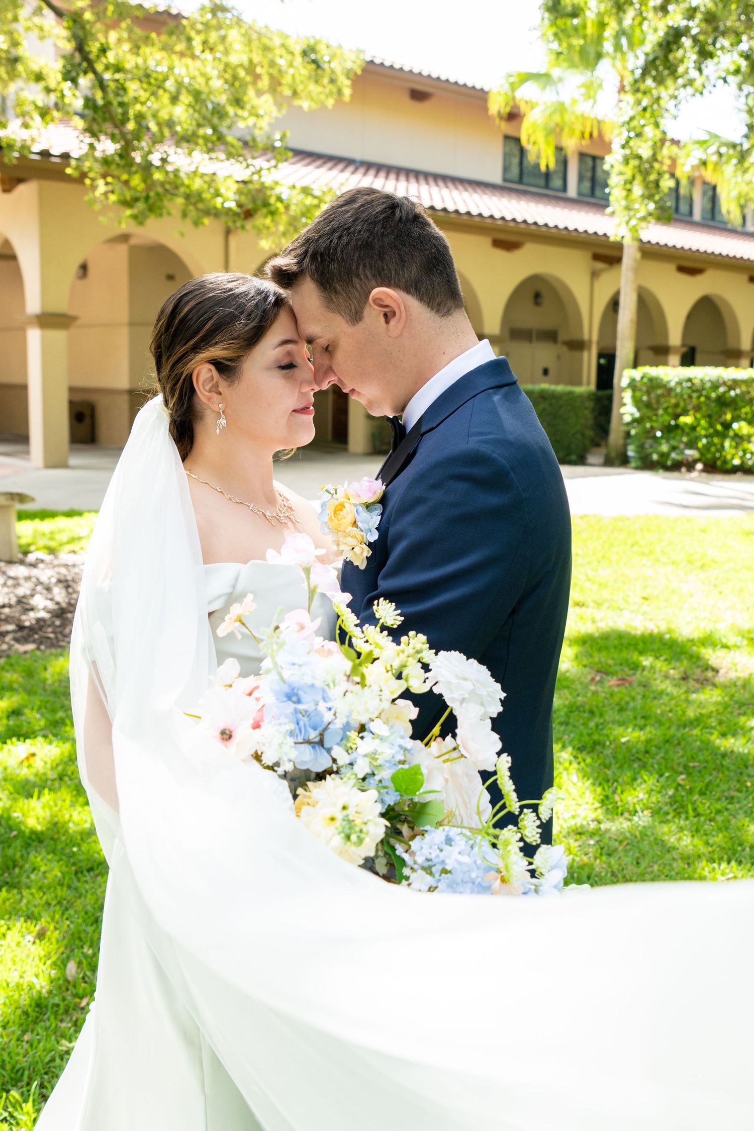 Bride and groom touching foreheads tenderly while holding a blue and white bouquet outdoors at a sunny wedding venue.