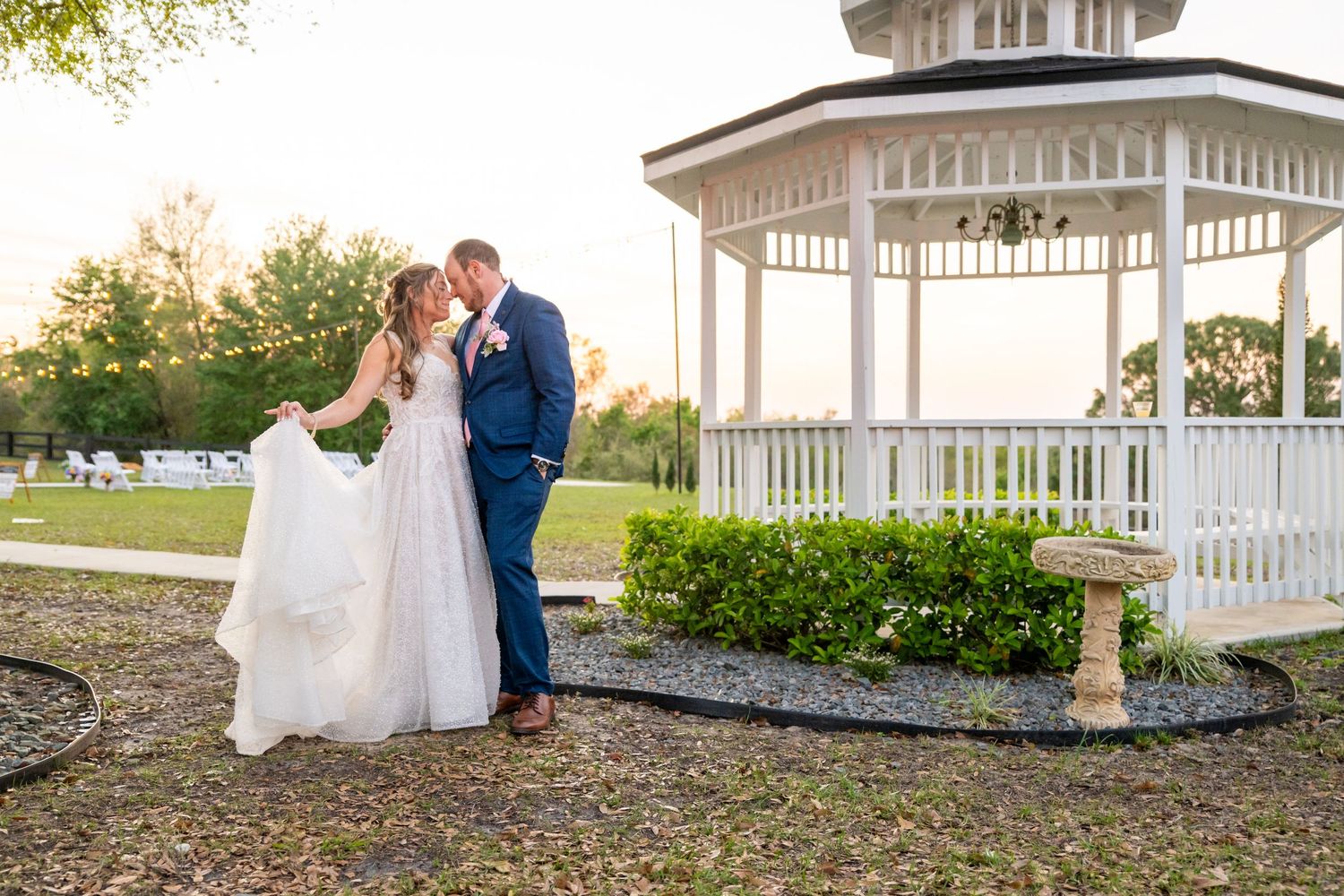 Bride and groom kiss near a white gazebo at sunset in a beautifully landscaped outdoor venue.
