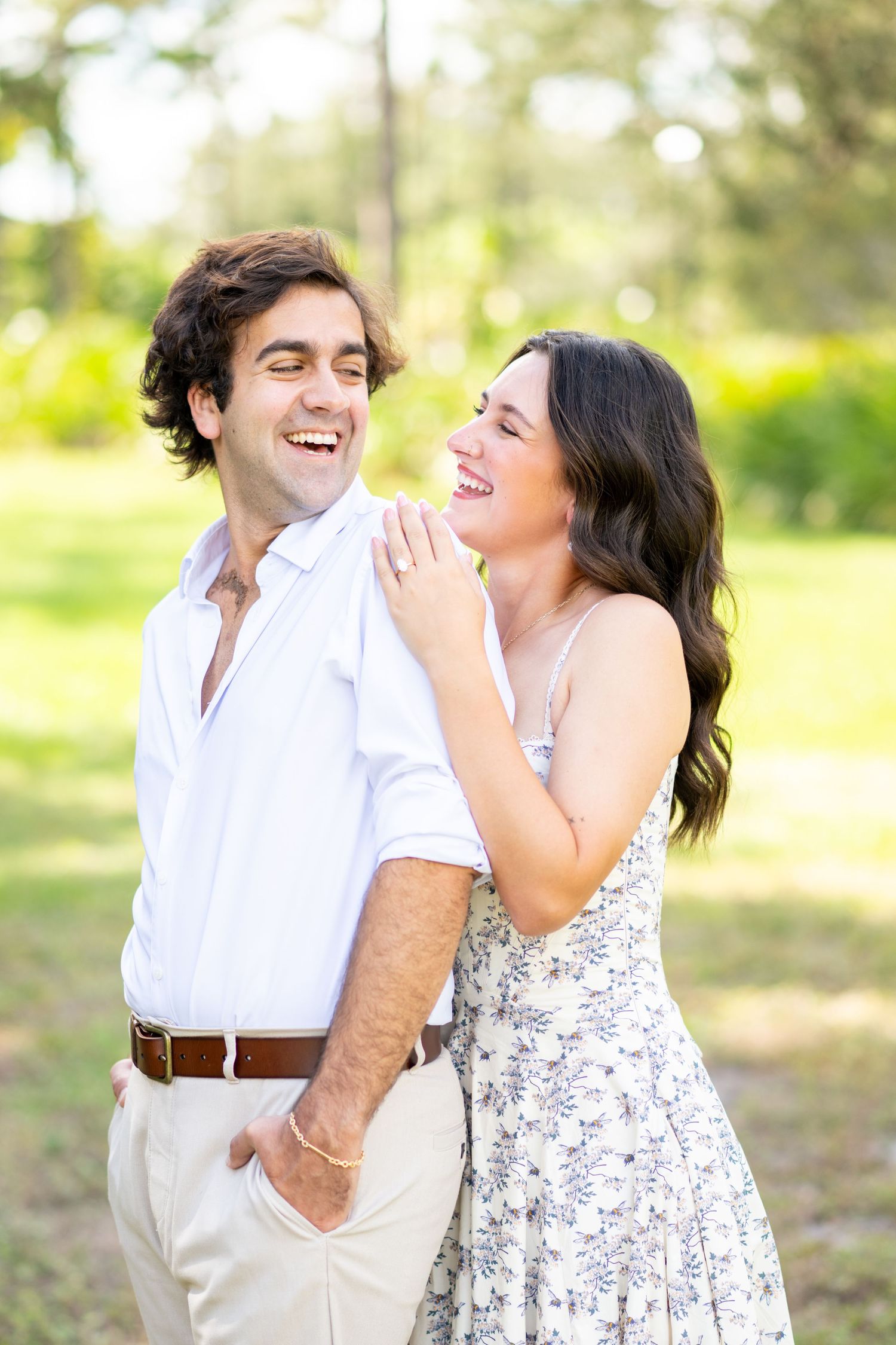 Joyful engaged couple laughing together outdoors in a sunlit green park setting.