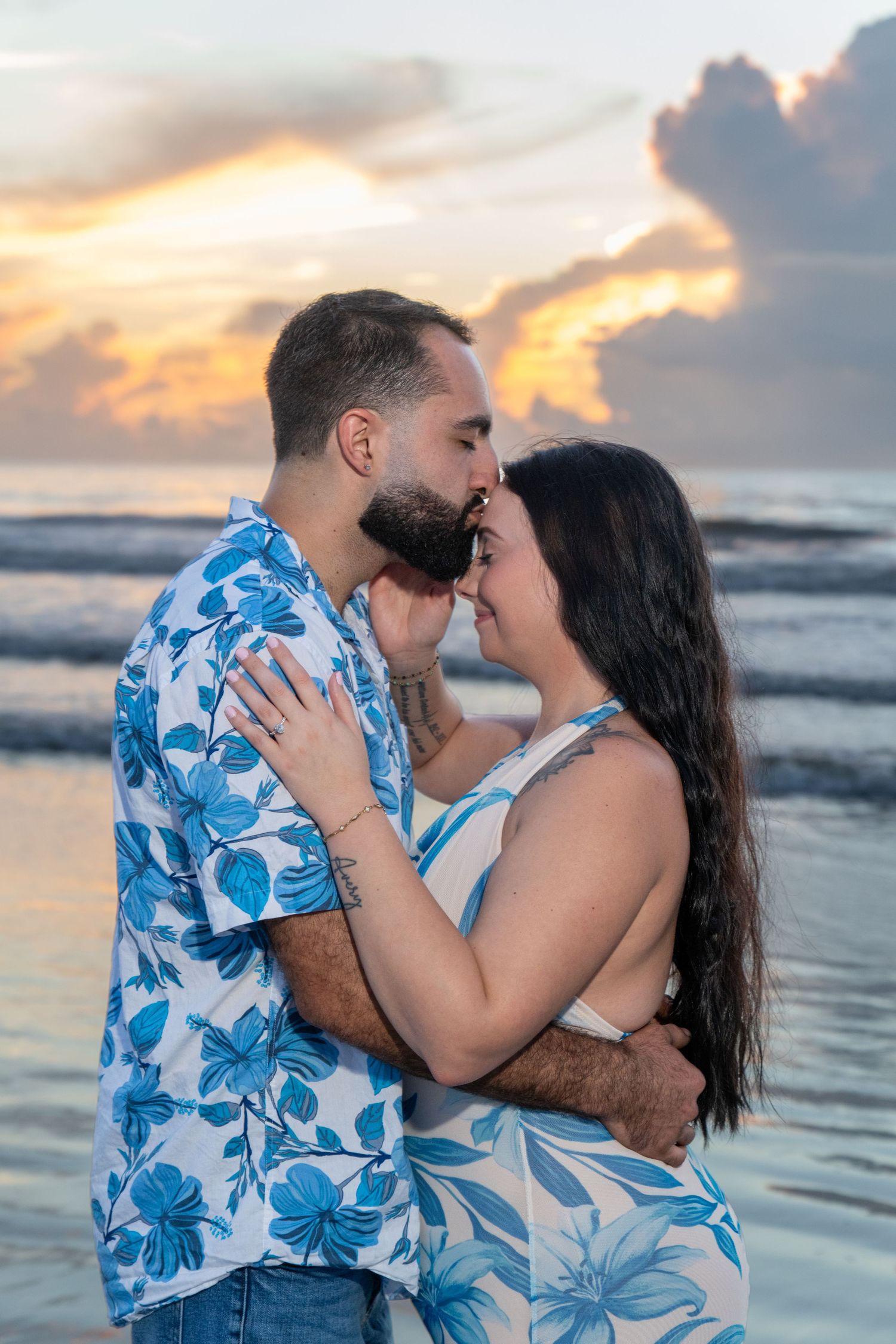Couple embracing and kissing at beach sunset, wearing matching blue floral outfits with ocean waves behind them.