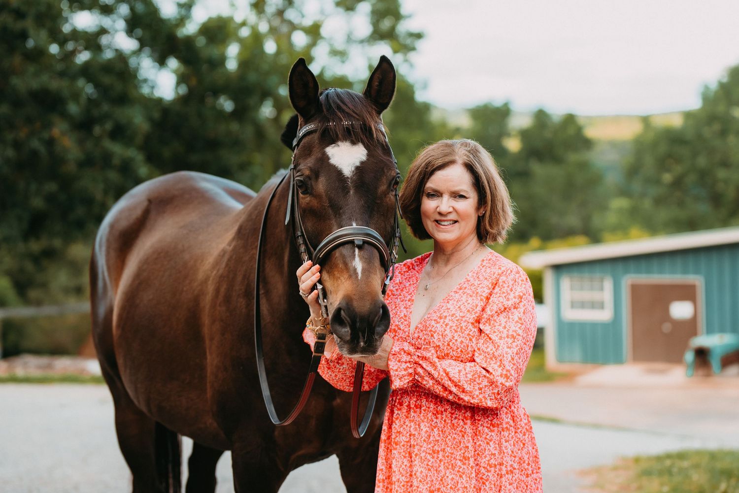Woman in red floral dress standing with a dark bay horse with a white star marking at an outdoor stable.