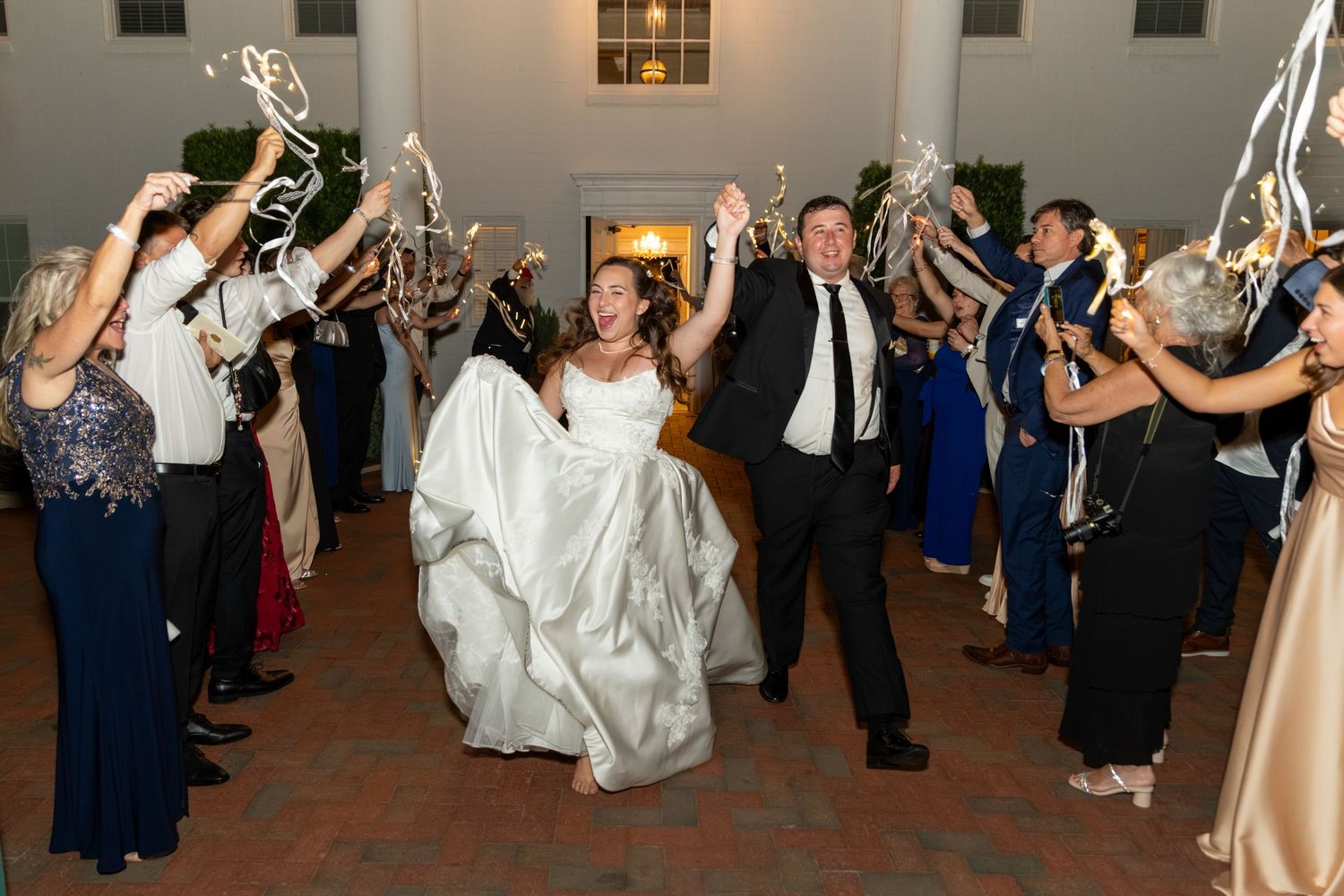 Bride and groom exit wedding reception through cheering guests waving ribbon wands in elegant venue.