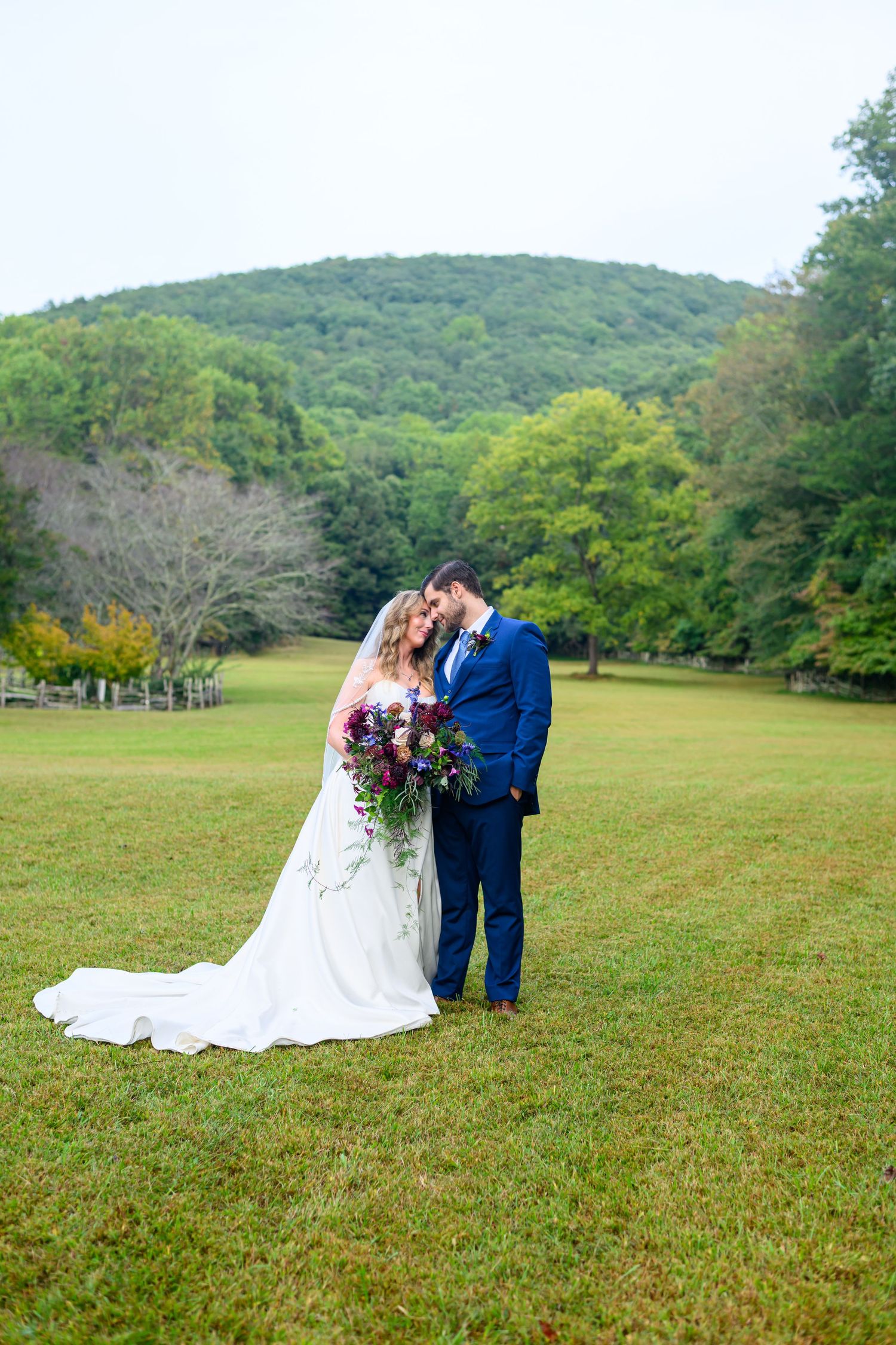 Bride and groom share a romantic forehead touch in a lush green meadow with rolling wooded hills backdrop.